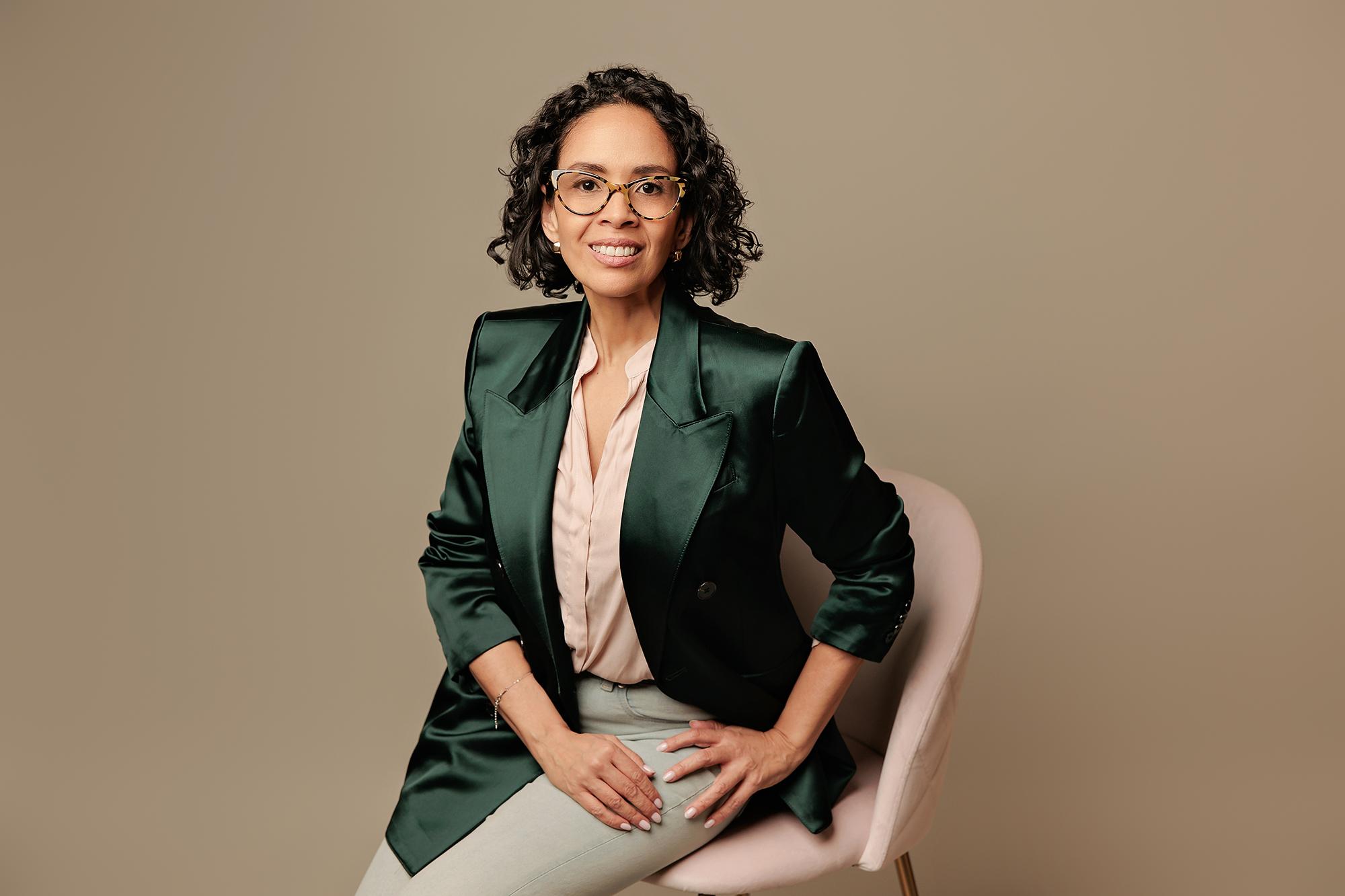 Calgary executive headshots: Smiling businesswoman with curly hair and tortoiseshell glasses wearing a green satin blazer, sitting on a pink chair in a photography studio