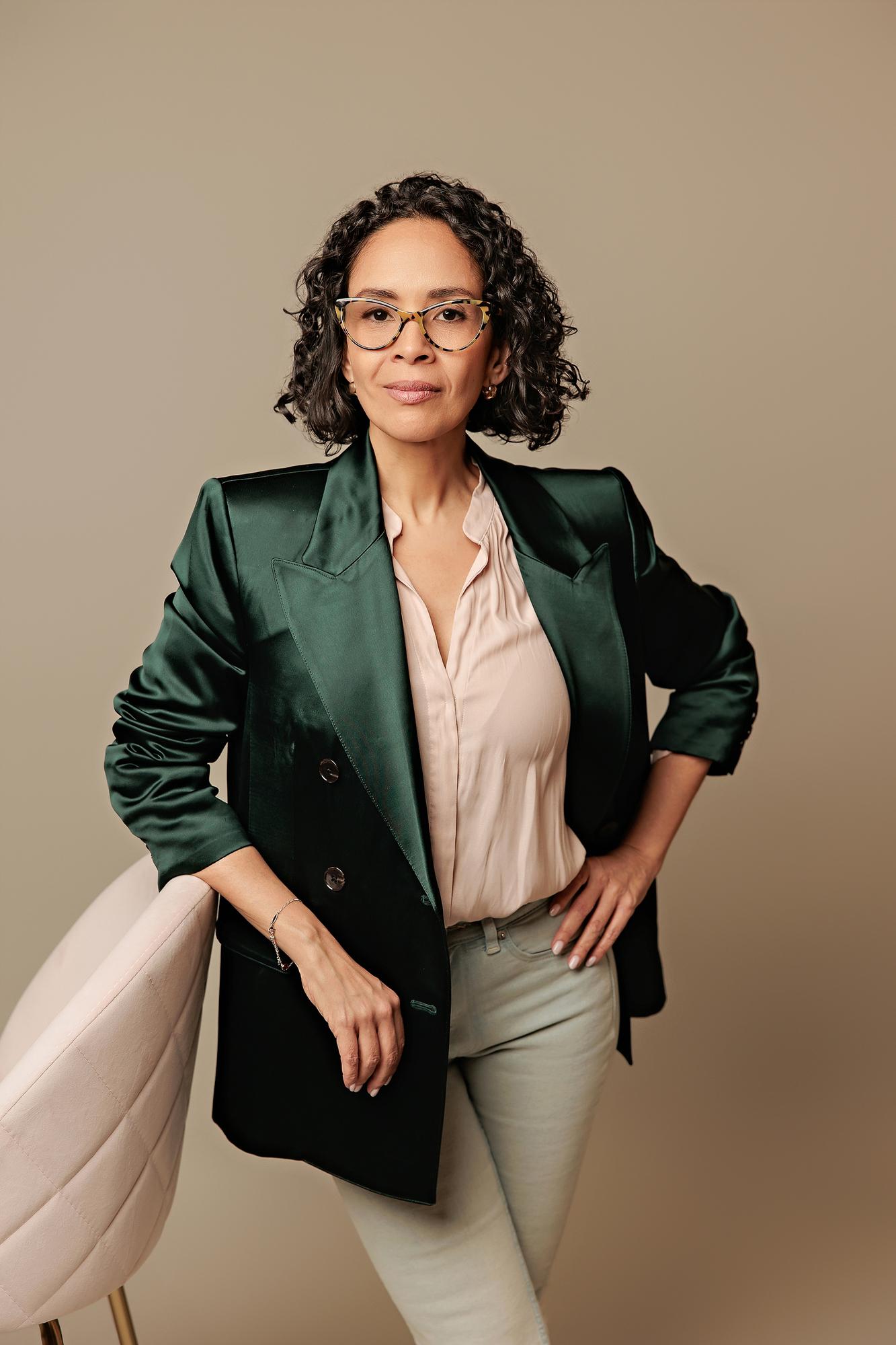Creative personal branding Calgary: Female professional in a green satin blazer and glasses leaning confidently against a pink velvet chair on a studio backdrop