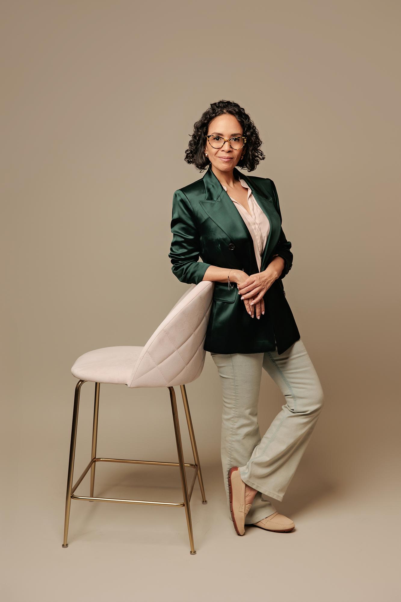 Personal branding photographer in Calgary: Businesswoman in a green satin blazer leaning confidently on a pink velvet barstool against a beige studio backdrop