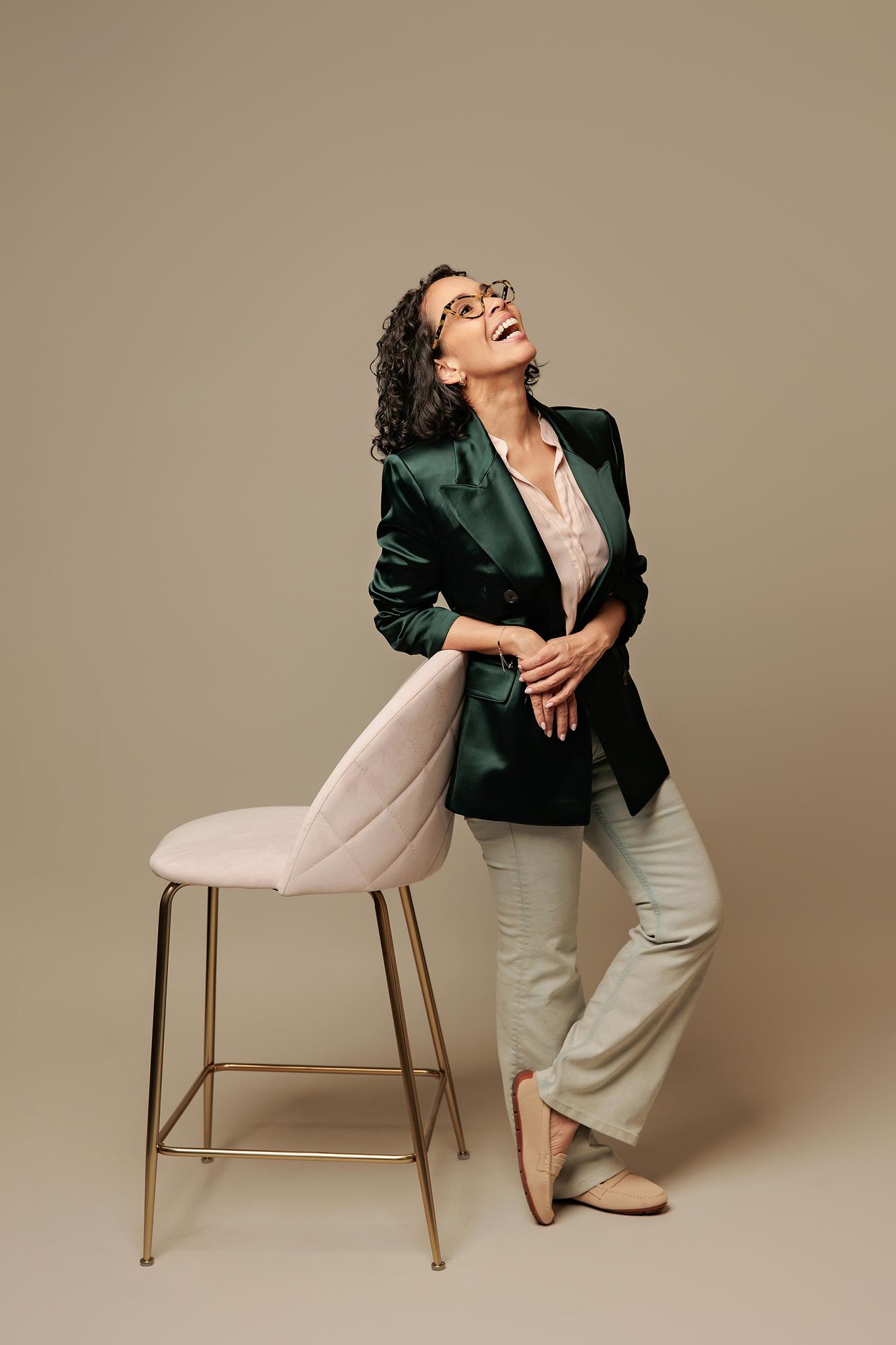 Calgary professional headshots: Joyful female entrepreneur laughing and looking up while leaning on a pink stool, wearing a green blazer in a photography studio