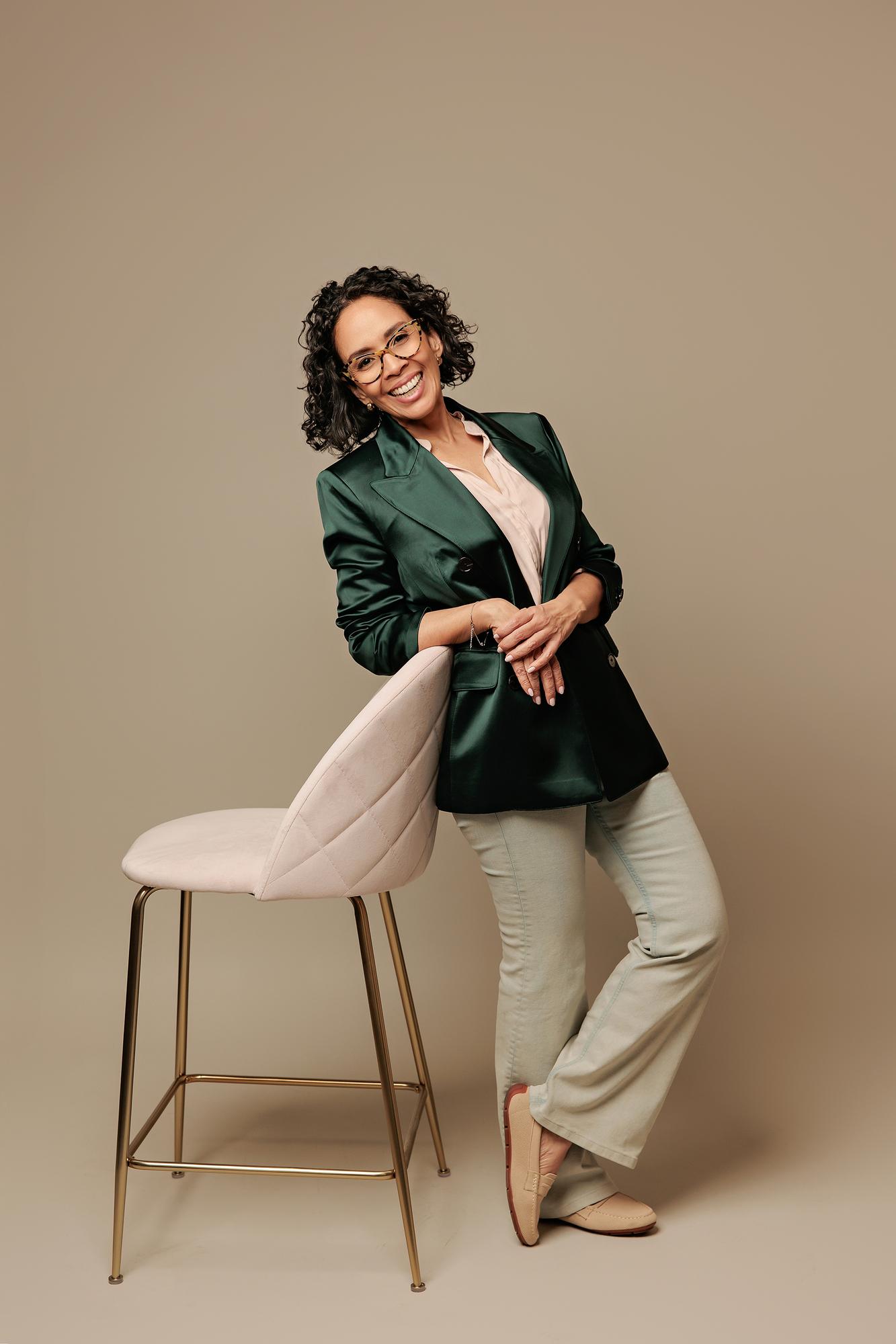 Corporate portrait photography Calgary: Smiling woman with glasses and a dark green blazer leaning on a pink chair against a seamless beige background