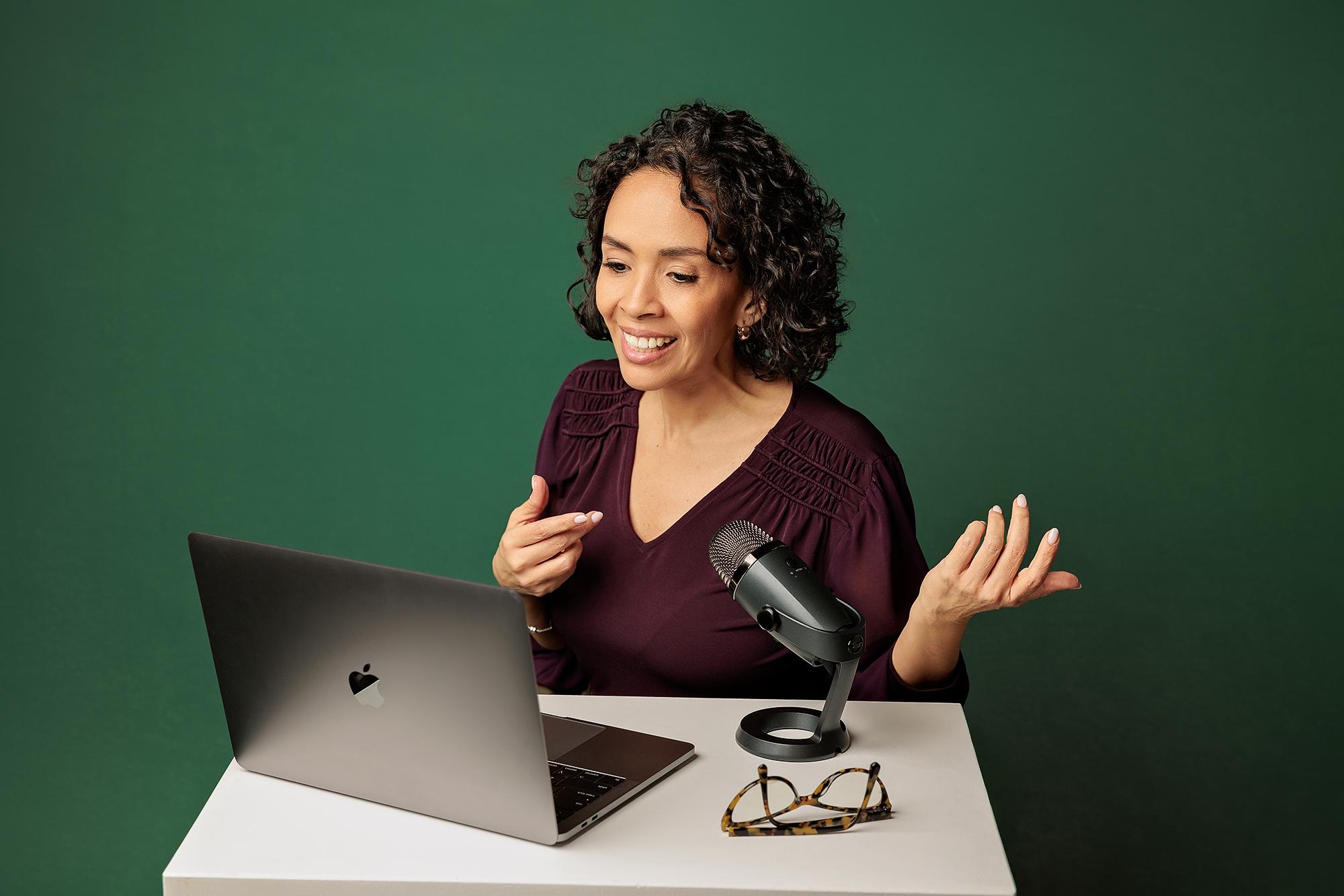 Podcaster branding photos Calgary: Female professional gesturing while speaking into a microphone during a laptop video call on a solid green backdrop