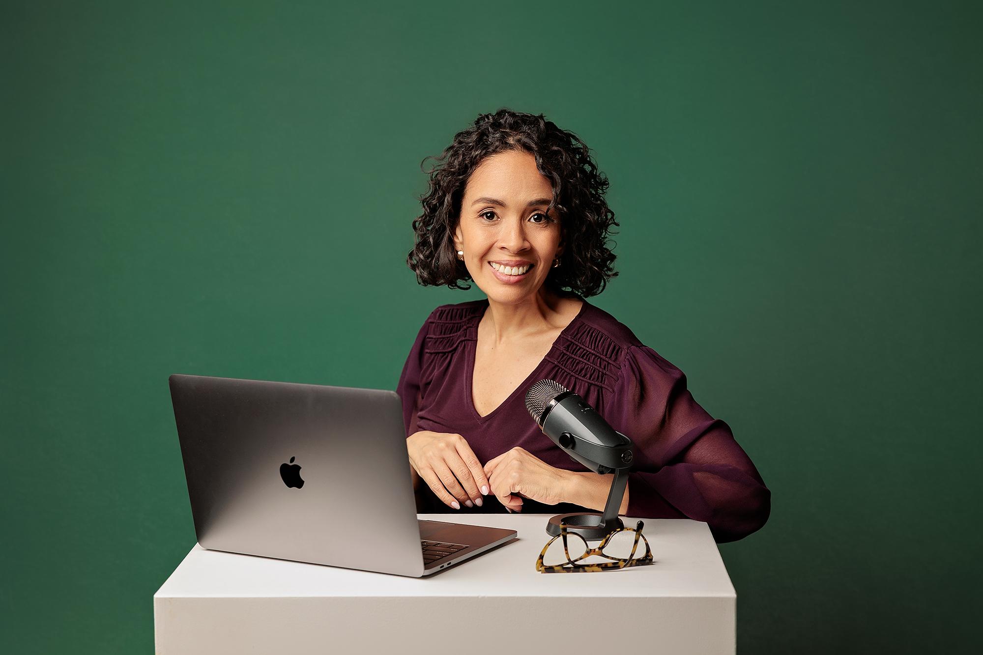 Calgary personal branding studio: Smiling woman in a plum blouse sitting at a white desk with a silver laptop and podcast microphone against a green background