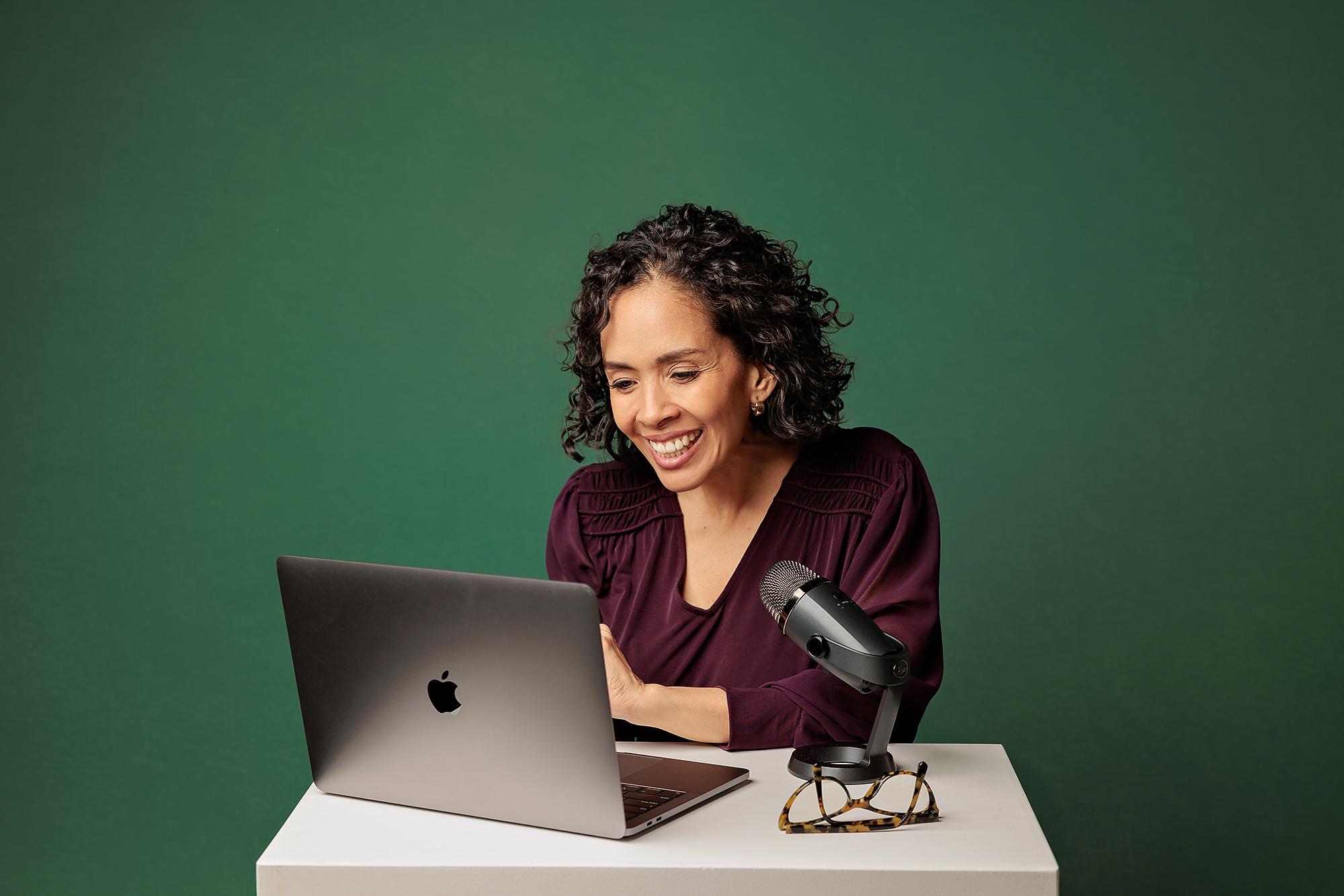 Professional headshot Calgary: Cheerful businesswoman laughing while looking at her laptop screen with a microphone on the desk, green studio backdrop