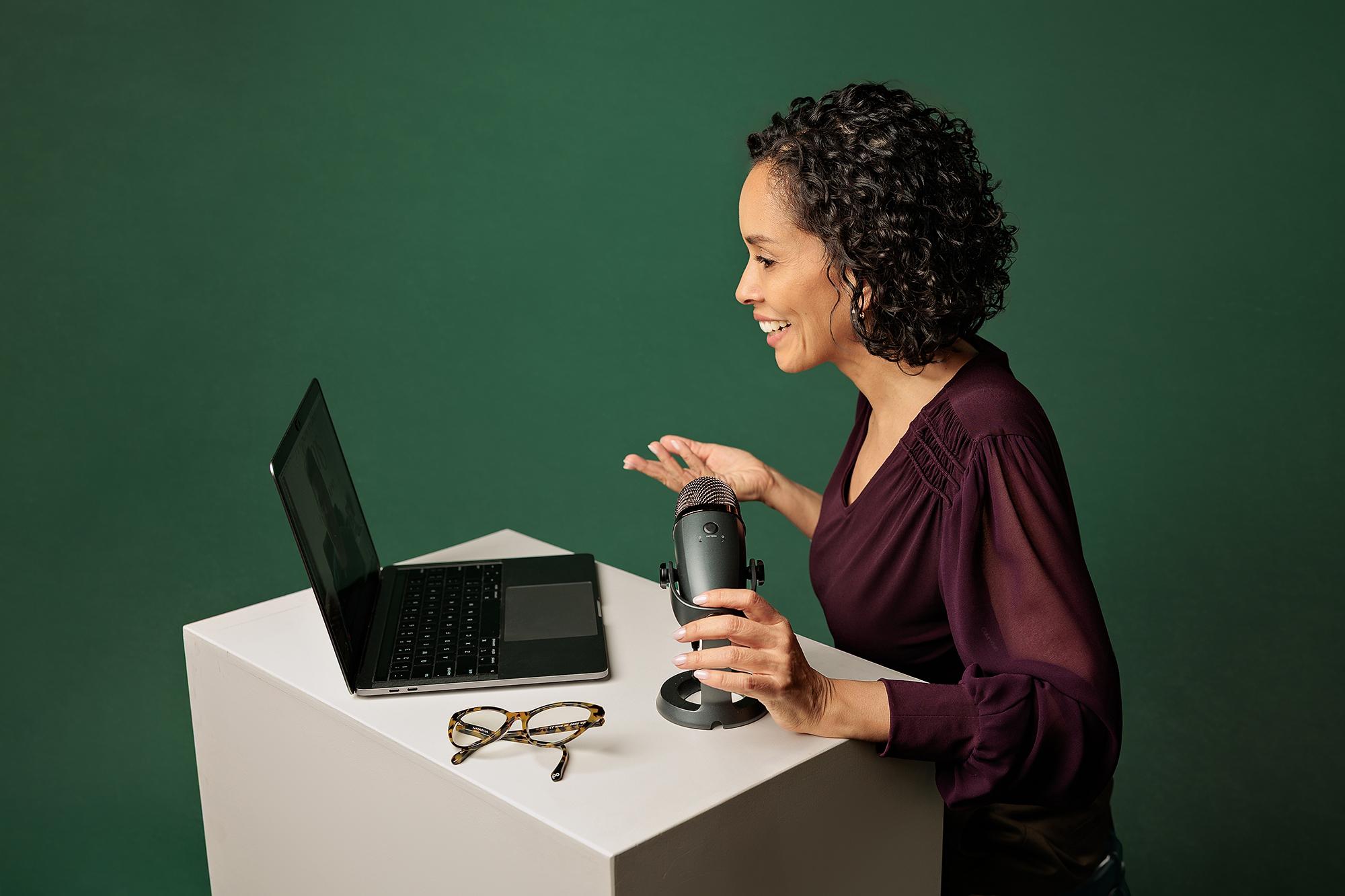 Business lifestyle photography Calgary: Female entrepreneur smiling and talking into a podcast microphone while looking at her laptop on a green background
