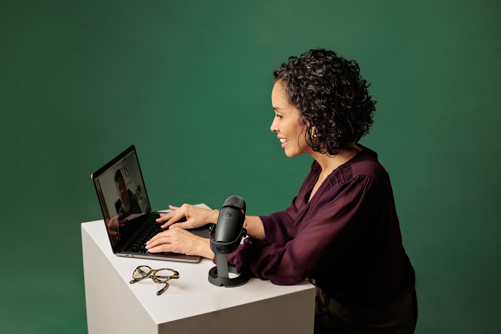 Calgary branding portraits: Professional woman typing on a silver laptop next to a studio microphone, wearing a burgundy blouse against a dark green background