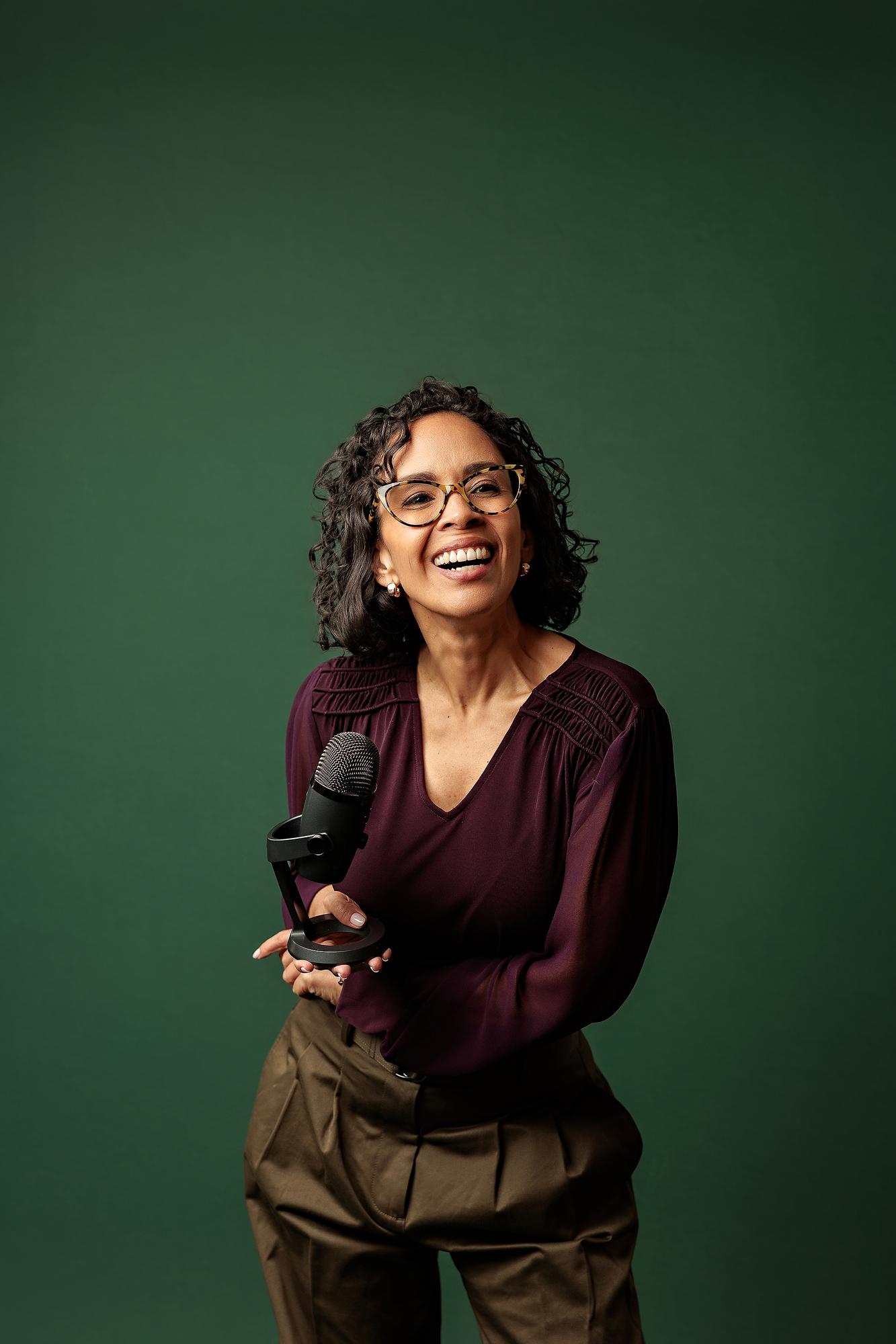 Creative personal branding Calgary: Joyful female podcaster laughing while standing and holding a microphone against a dark green studio backdrop