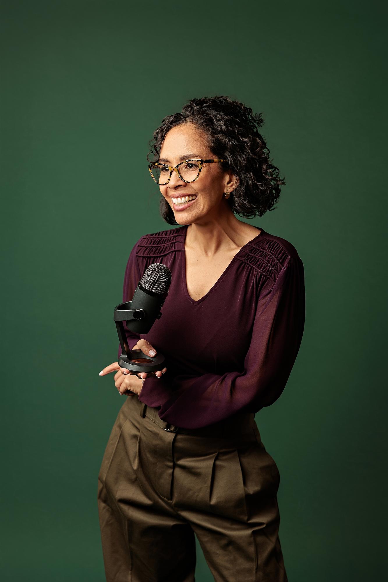 Calgary headshot photographer: Smiling businesswoman wearing a burgundy blouse and glasses, holding a podcast microphone against a solid green background