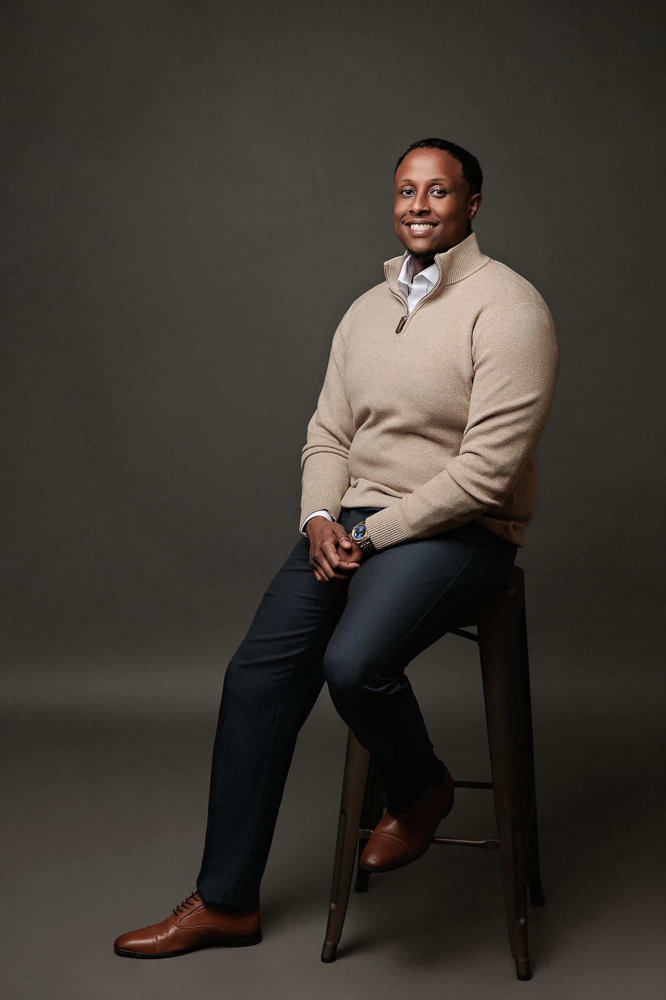 A smiling man in a beige sweater sits on a stool for a professional headshot at our Calgary photography studio, featuring soft lighting and a dark backdrop.