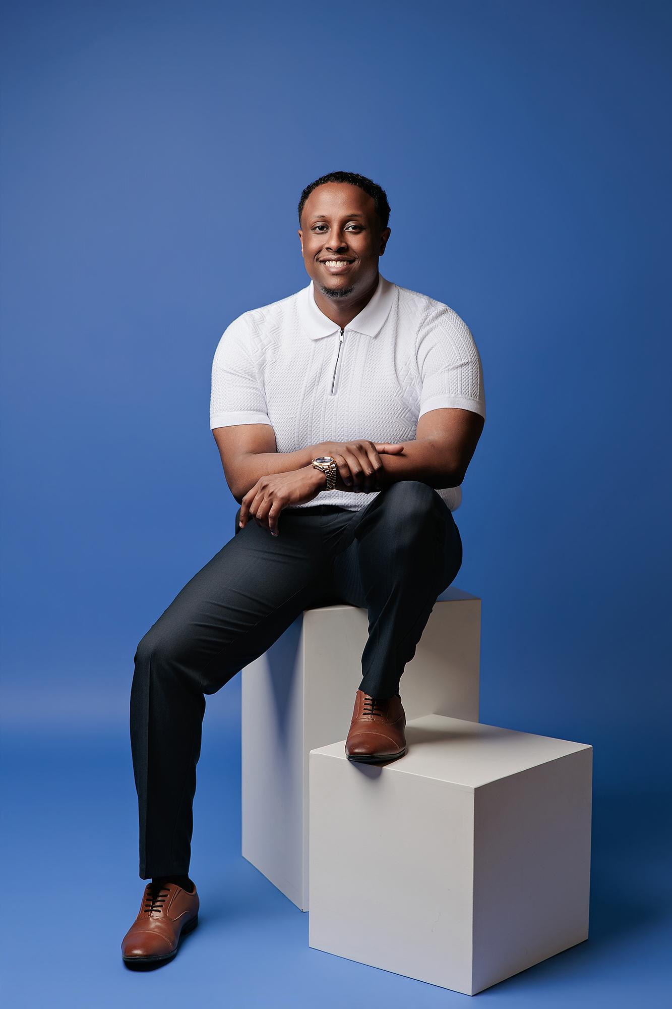 A smiling man in a white polo shirt sits on white studio blocks, showcasing the modern style of our Calgary headshot photography studio.