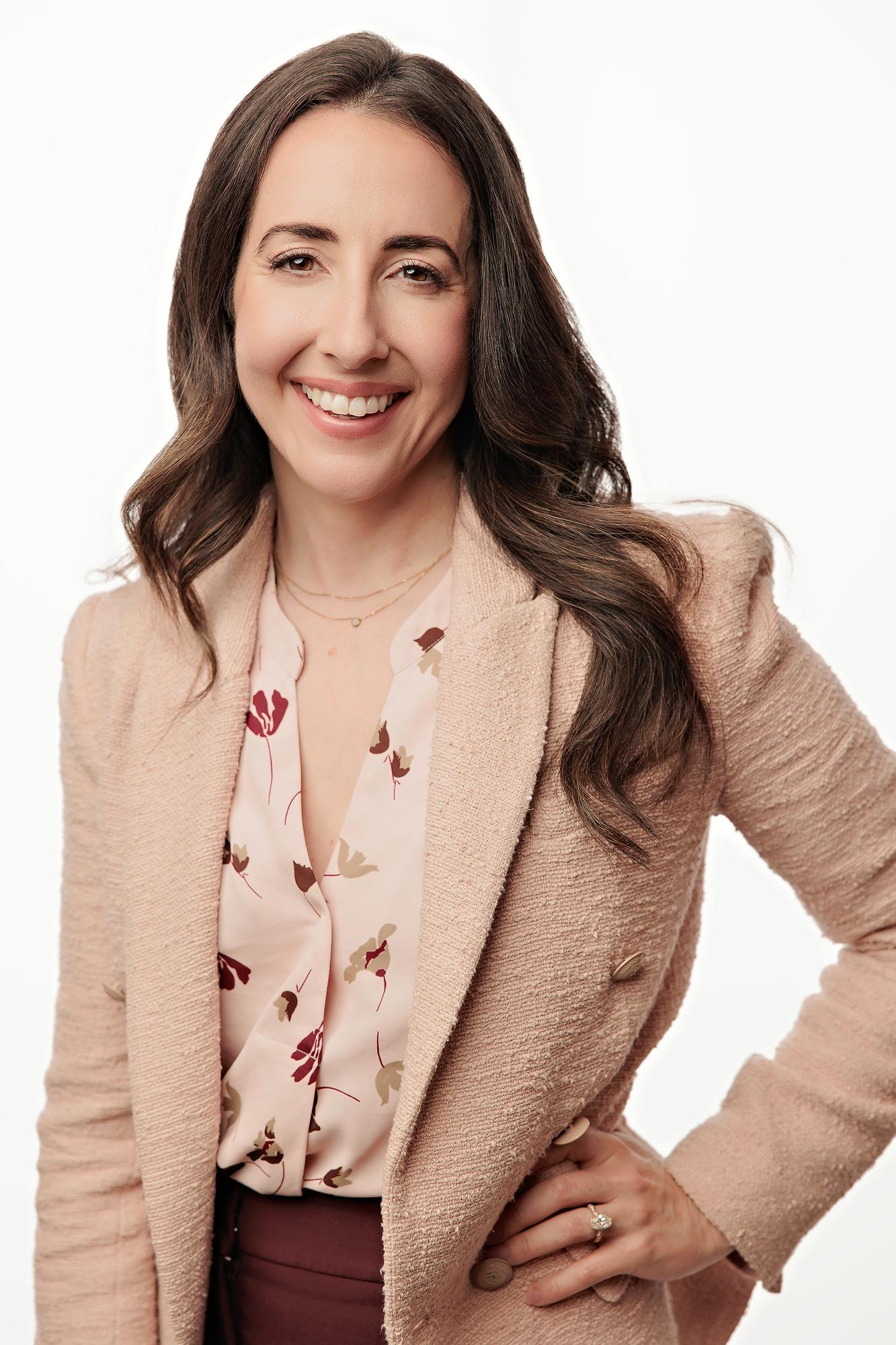 Professional headshot Calgary: Smiling woman with dark wavy hair wearing a textured pink blazer over a floral blouse against a clean white background