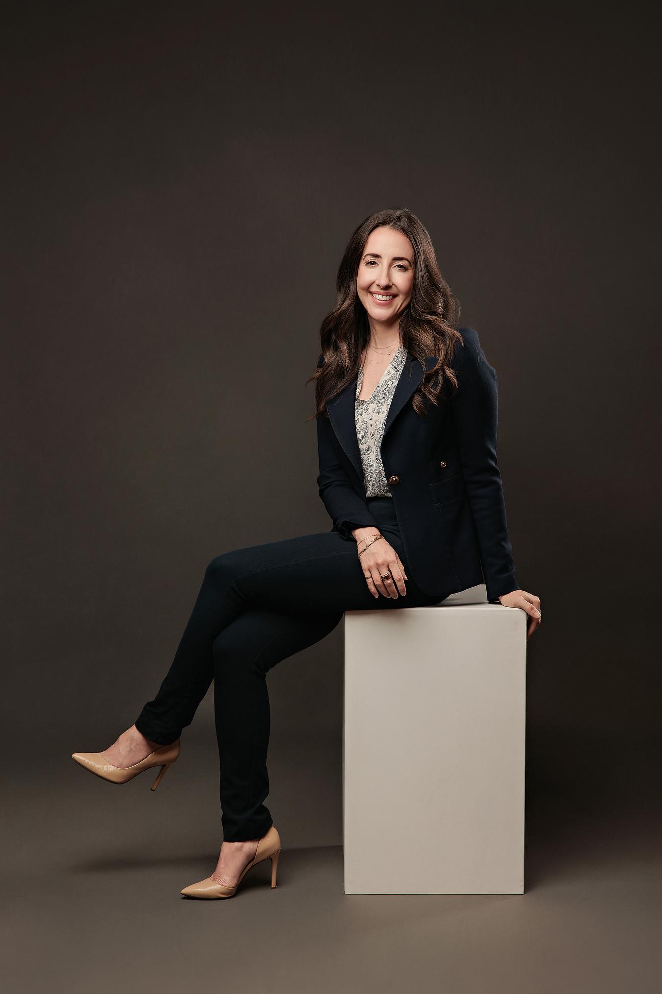 Executive headshot photography Calgary: Professional woman in navy blazer leaning casually on a white studio cube against a dark background
