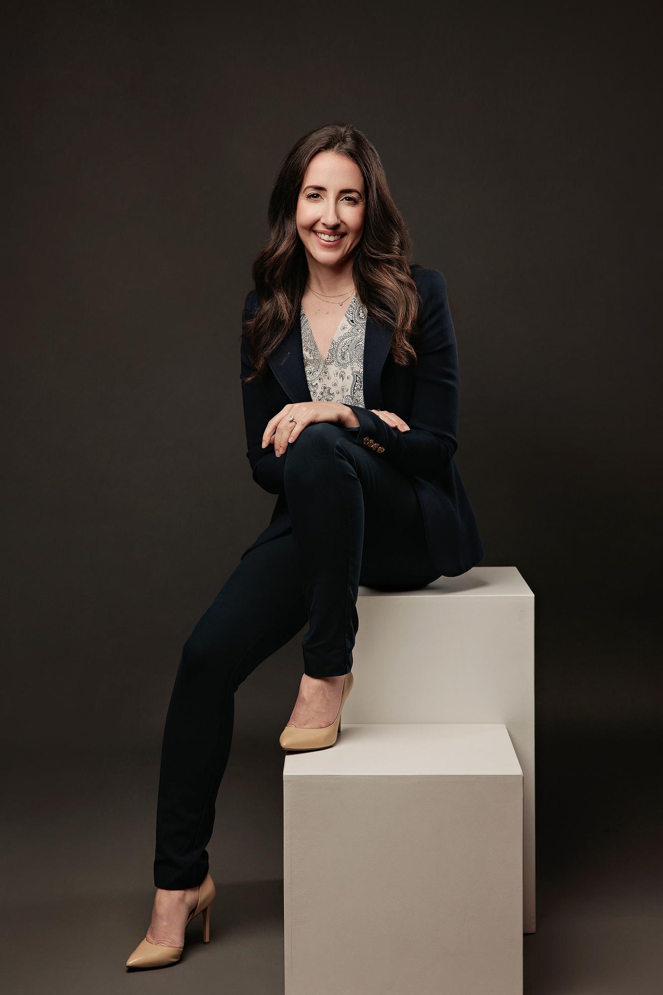 Business headshot photography Calgary studio: Professional woman in navy blazer seated on a two-level white cube leaning forward confidently