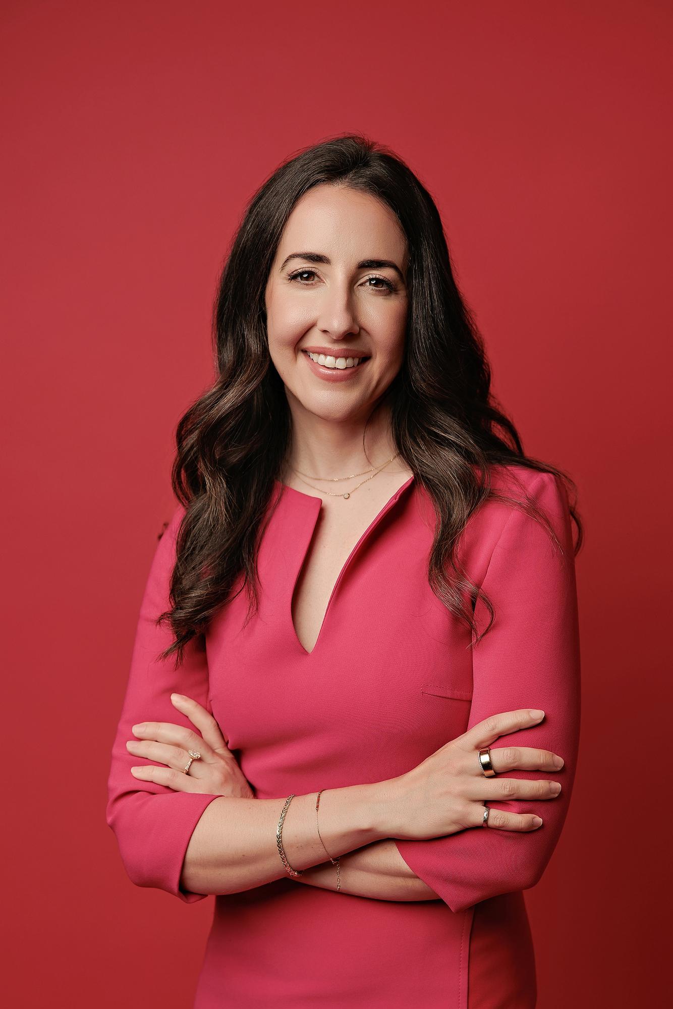 Corporate headshots Calgary: Confident businesswoman wearing a bright pink dress standing with arms crossed against a seamless red studio backdrop