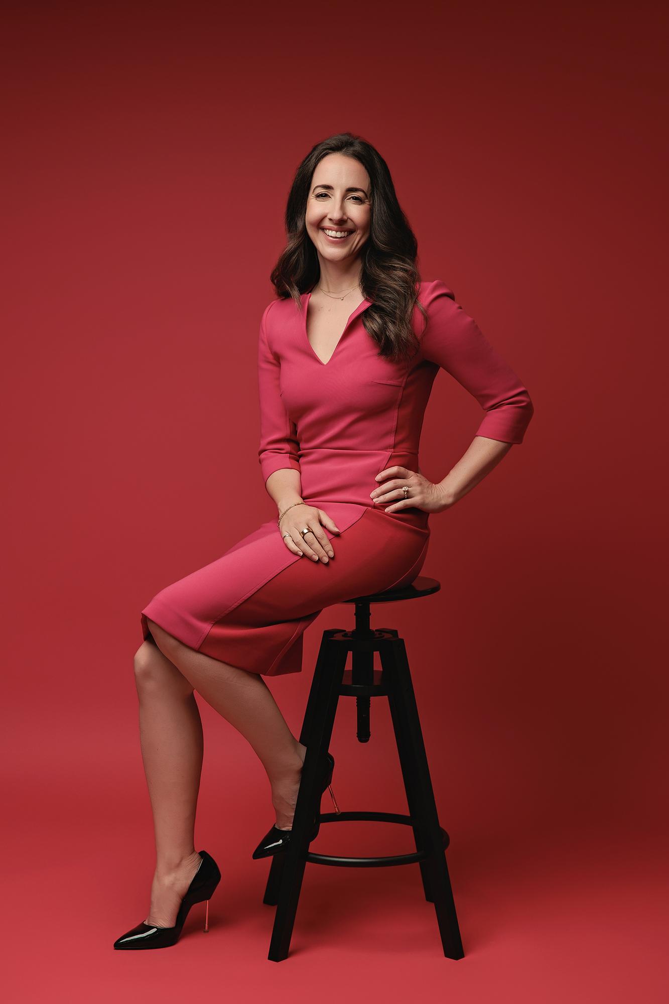 Best headshot photographer Calgary: Professional woman in a pink dress sitting on a black studio stool with one hand on her hip against a red backdrop
