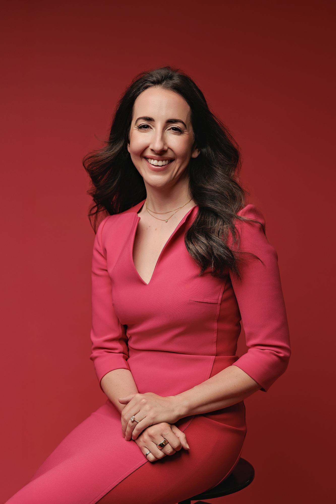 Premium headshot session Calgary: Joyful businesswoman sitting on a studio stool smiling warmly against a bold red background