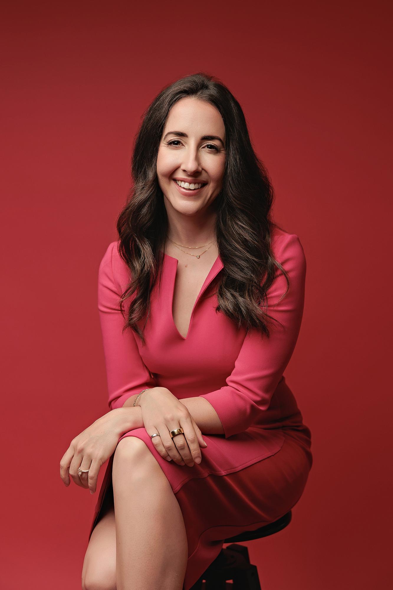 Headshot photography Calgary professional woman: Smiling corporate executive in a pink dress leaning forward with hands crossed on her knee, red studio background