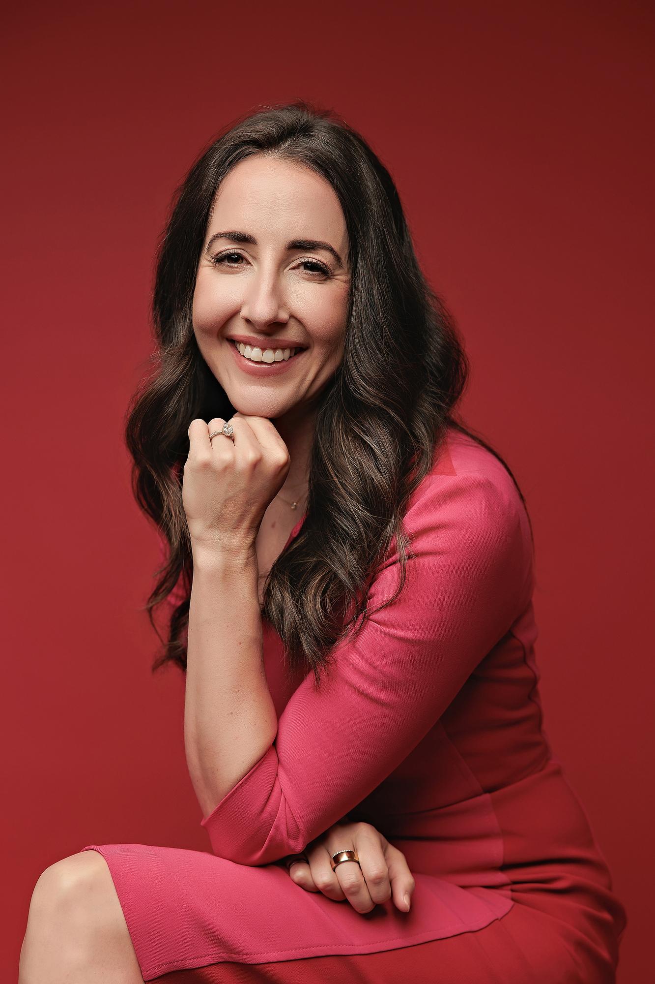 High quality headshots Calgary downtown studio: Beautiful professional woman in a bright pink dress resting her chin on her hand against a vivid red backdrop