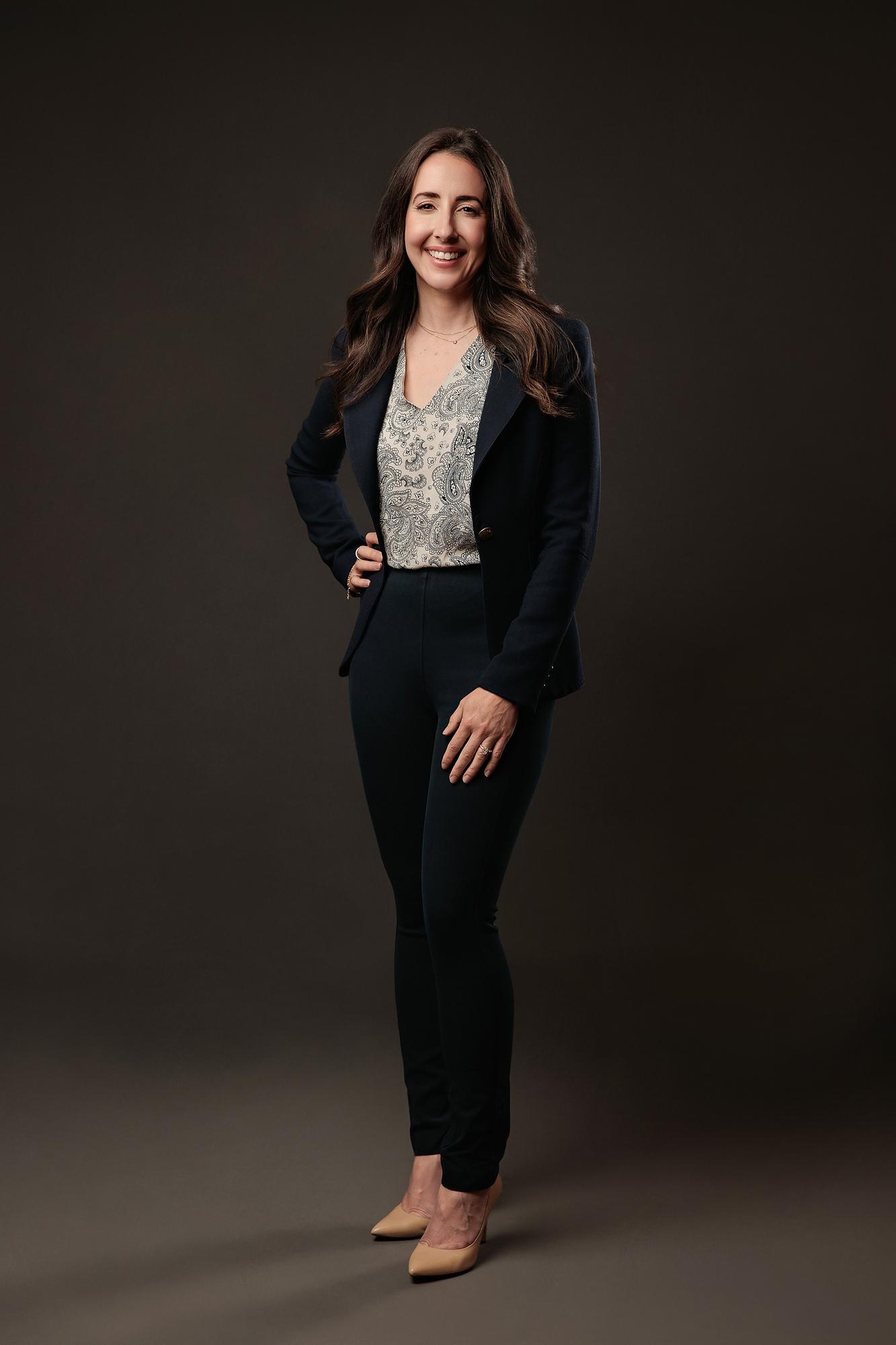 Professional headshot Calgary: Woman standing confidently with hand on hip wearing a navy blazer and paisley blouse against a dark studio background