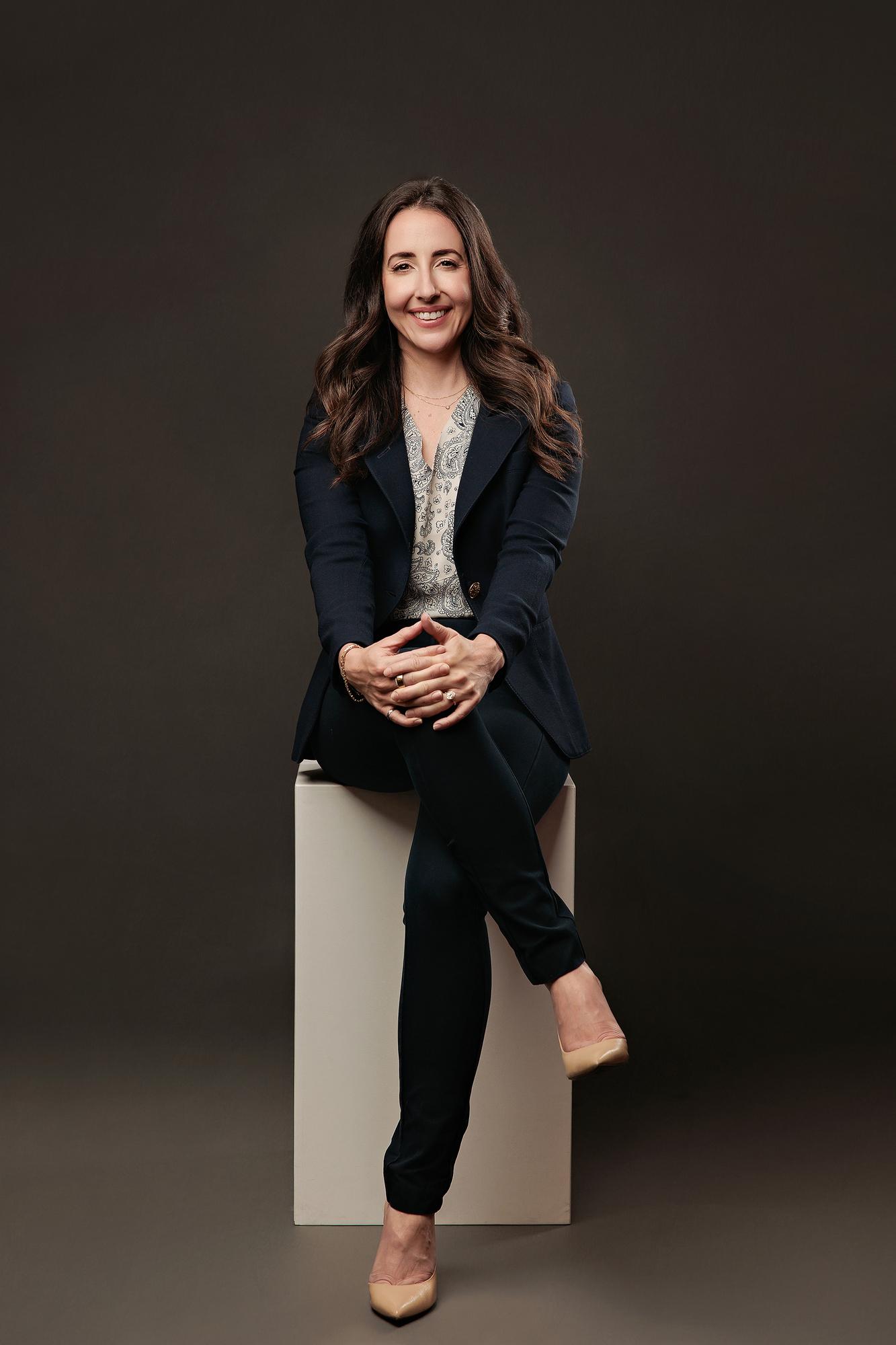 Corporate headshot Calgary studio: Professional woman in navy blazer seated on a white cube with hands clasped, smiling warmly against a dark background