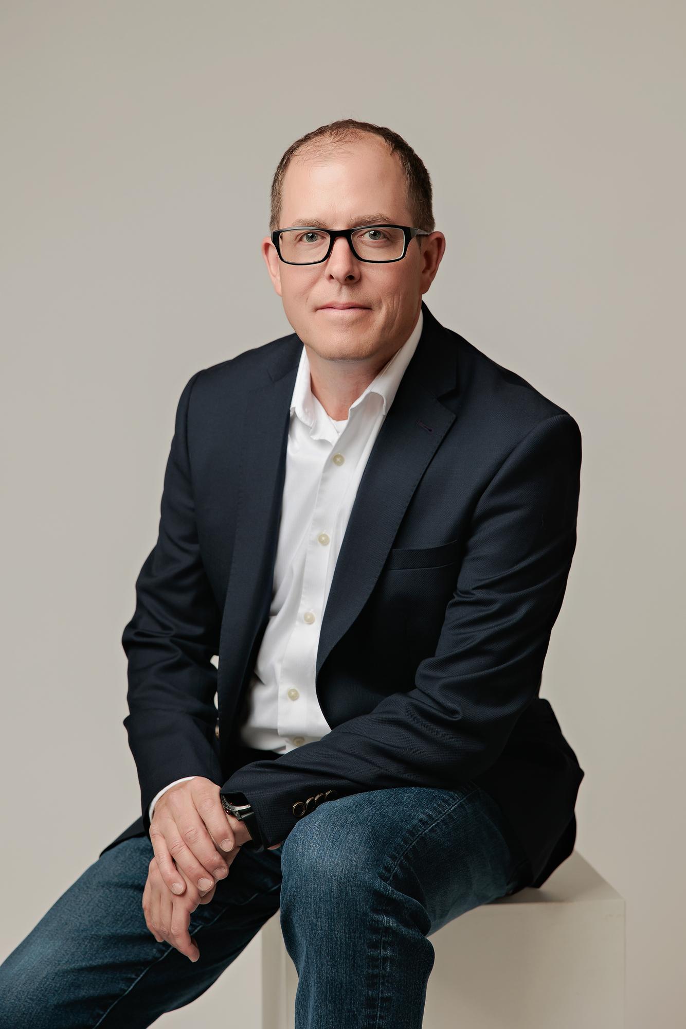 Professional headshot Calgary: Man in navy blazer and jeans seated on a white cube with hands relaxed, light studio background