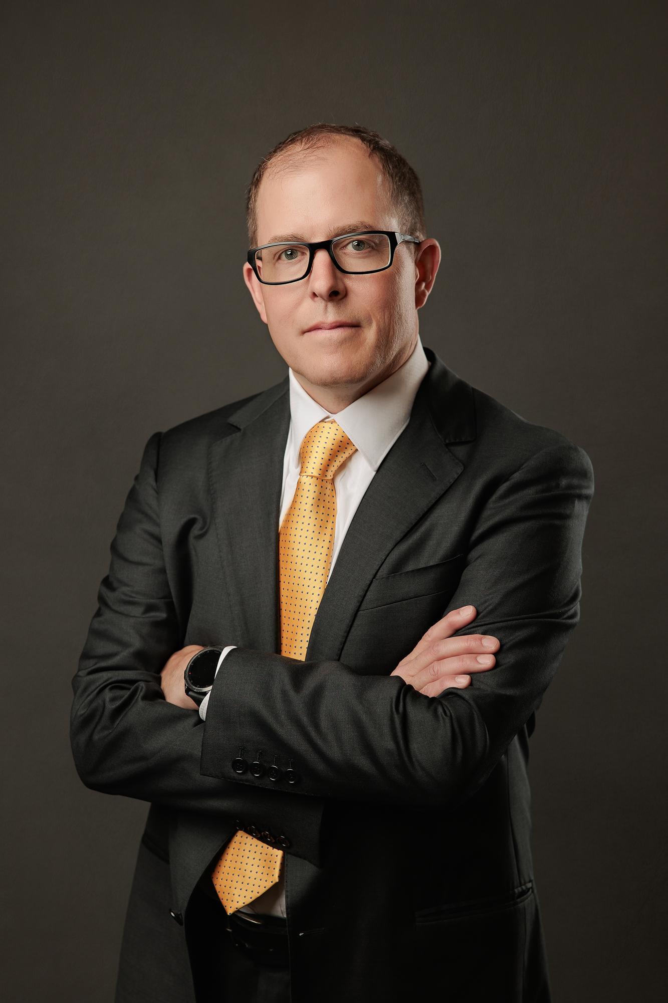 Executive headshot Calgary: Confident man in dark suit with gold tie standing with arms crossed against a dark studio background