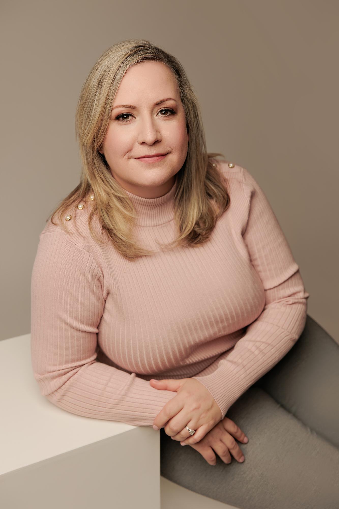 Professional headshot Calgary: Warm and approachable woman in a pink ribbed turtleneck leaning on a white surface against a neutral studio background
