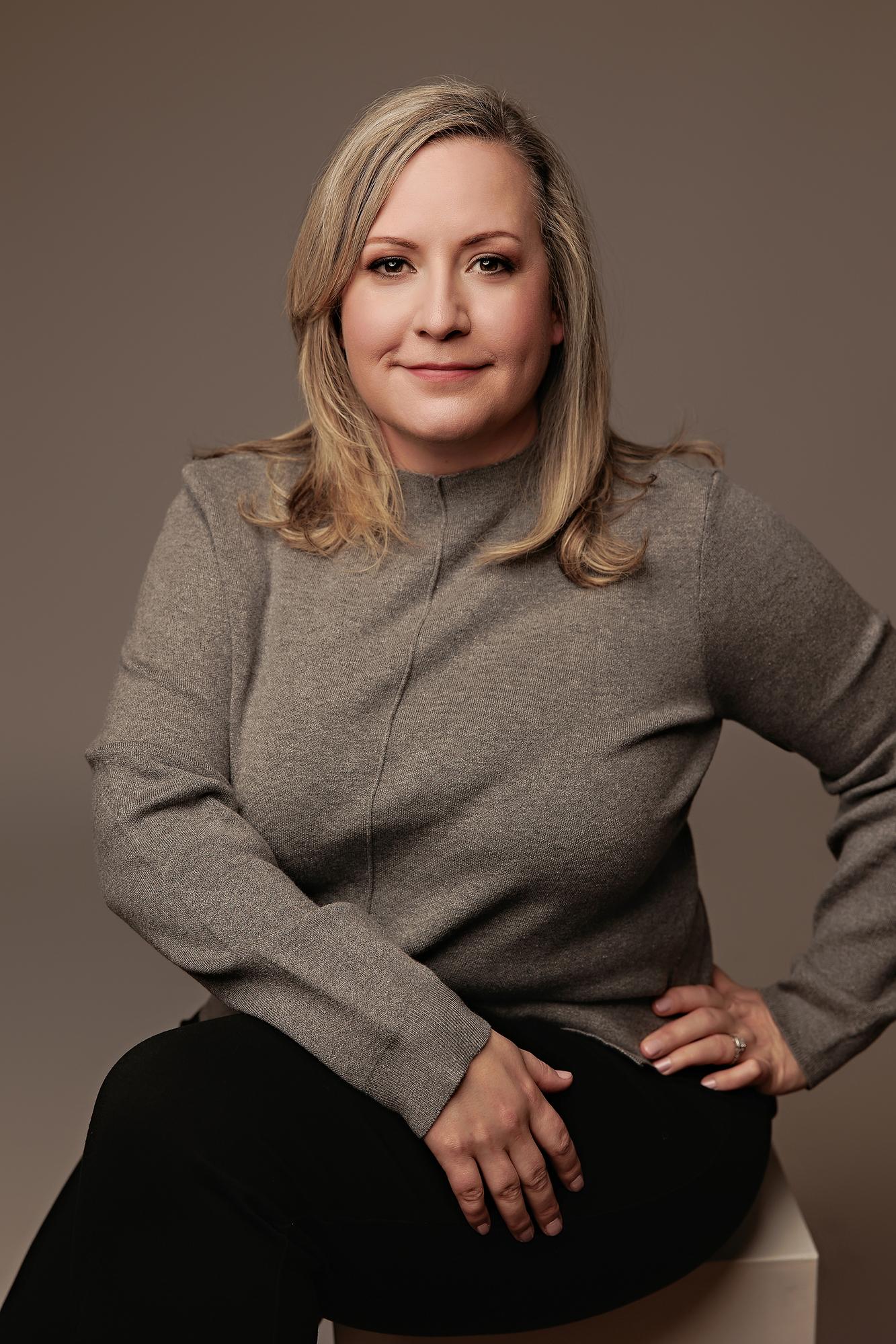 Calgary headshot photographer studio portrait: Professional woman in grey turtleneck sweater seated with hand on hip against a warm taupe background