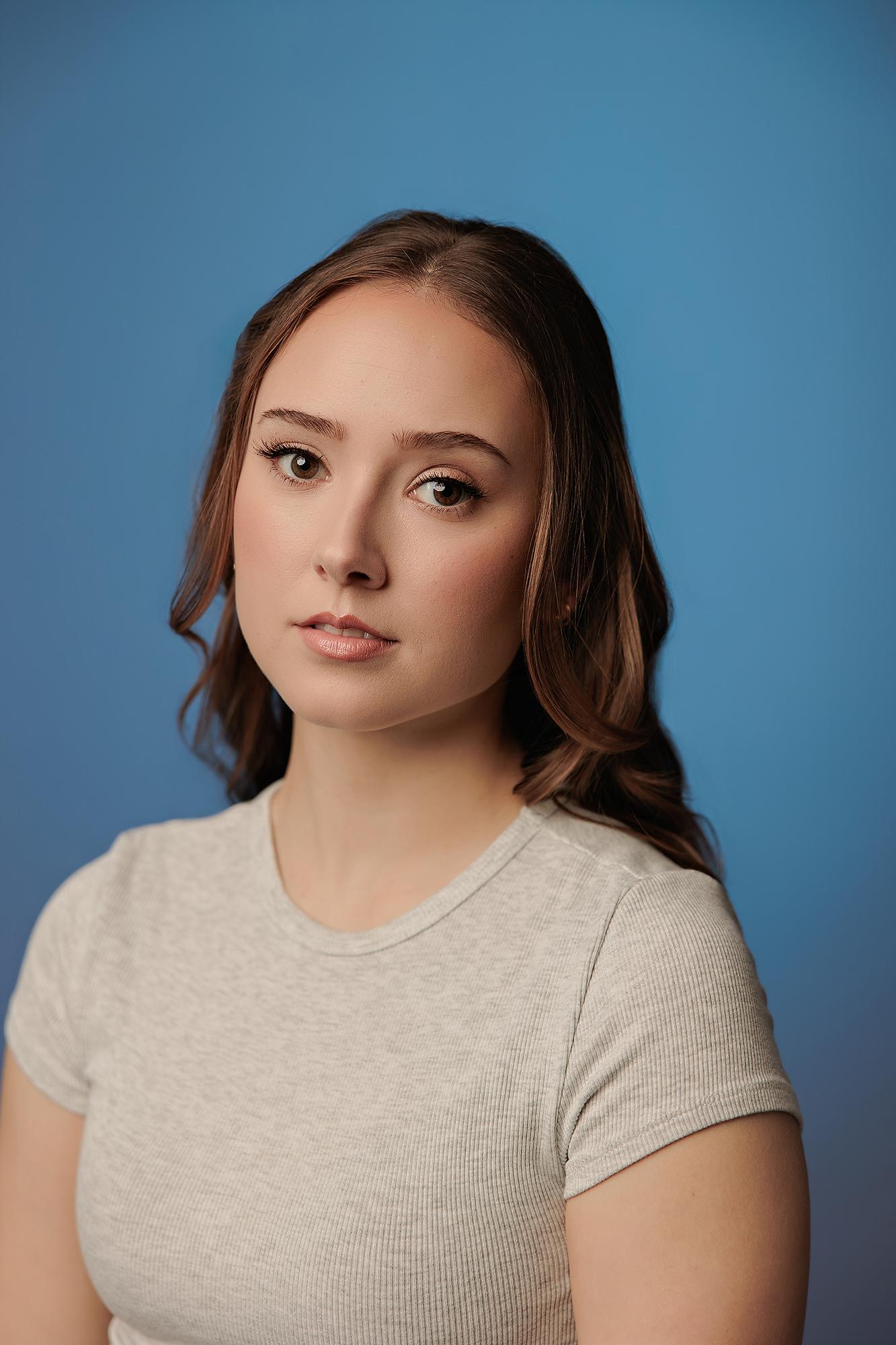 A young woman with soft brown hair and a neutral expression wears a grey ribbed shirt against a blue background, a polished professional headshot from Amanda Dams Headshots studio