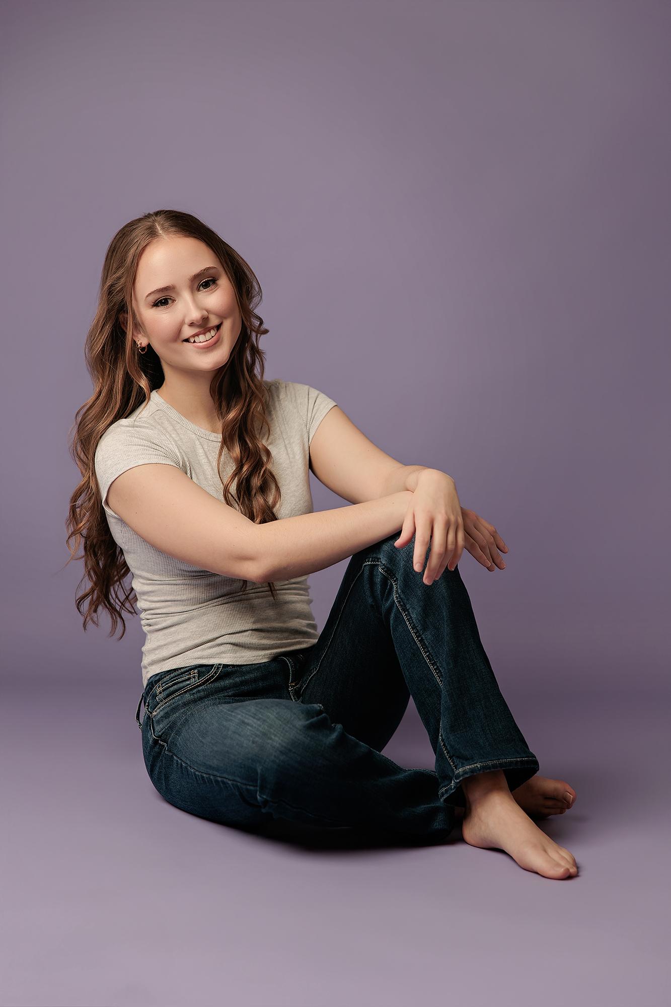 A smiling young woman with long wavy hair, in a grey tee and jeans, sits casually against a vibrant purple studio backdrop, perfect for Calgary personal bran...