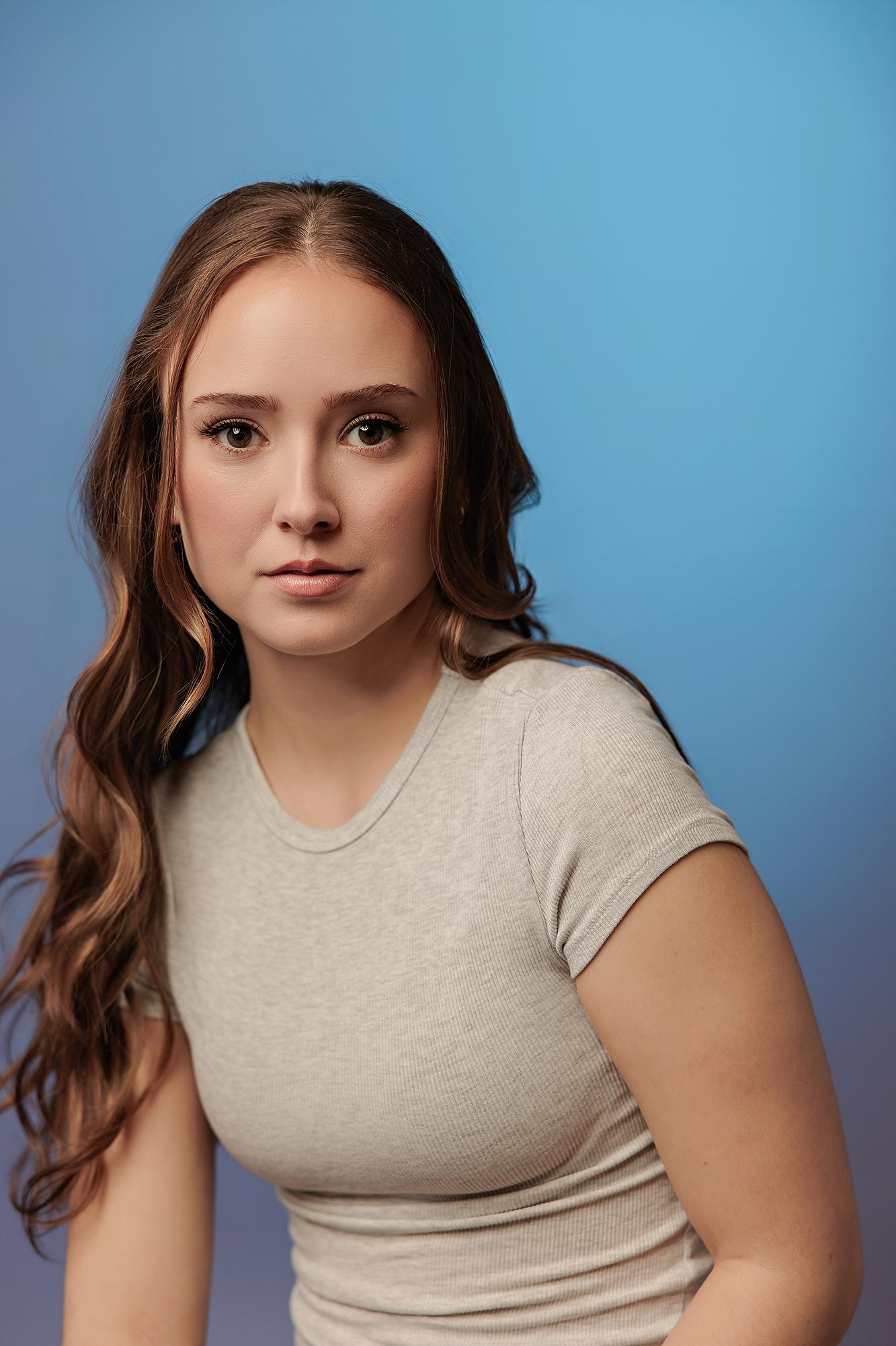 A young woman with wavy brown hair and a confident expression wears a grey t-shirt against a blue studio background, exemplifying a professional Calgary headshot