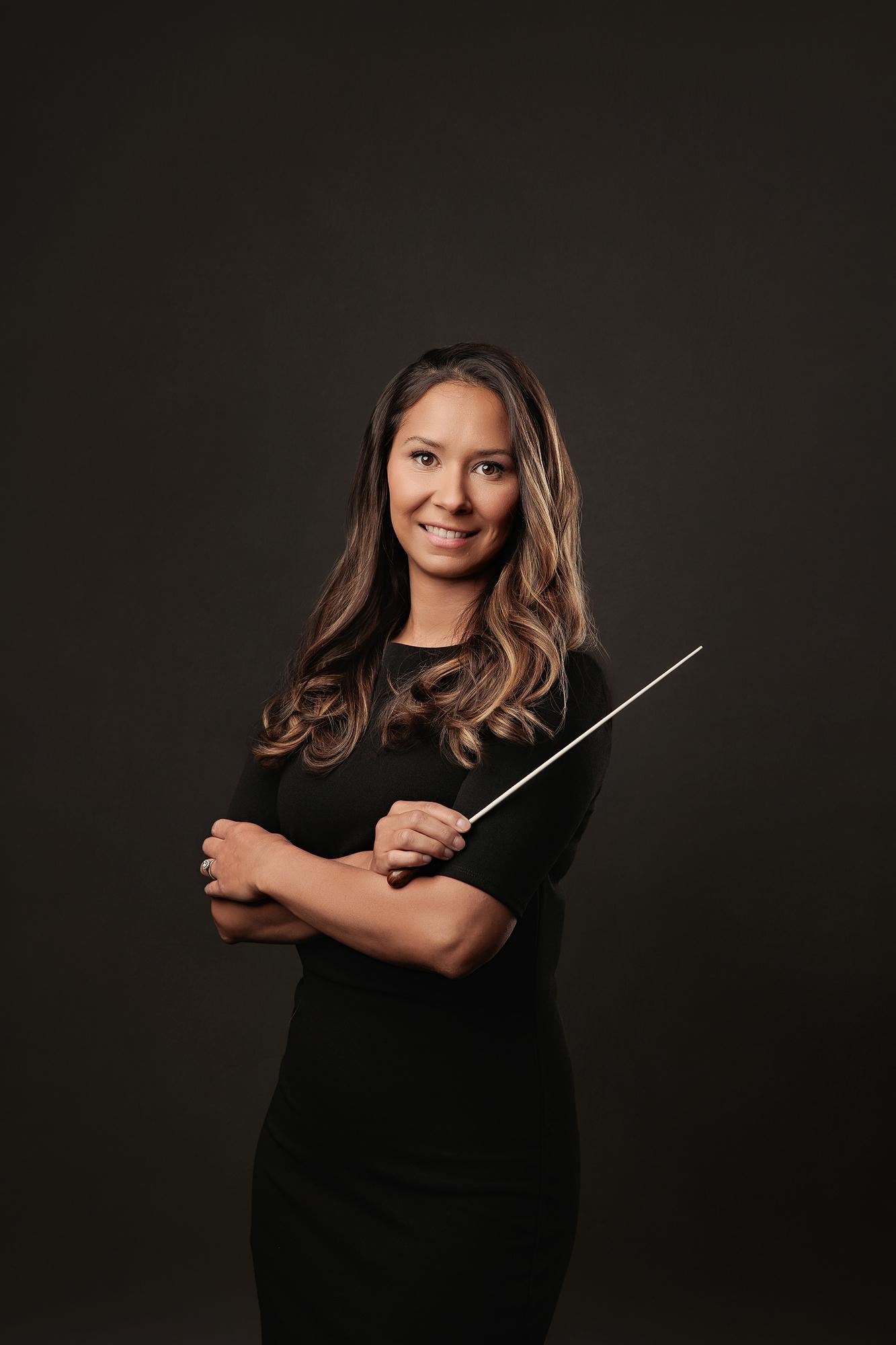 Creative professional headshot Calgary: Female conductor in a black dress standing with arms crossed holding a baton against a dark studio background
