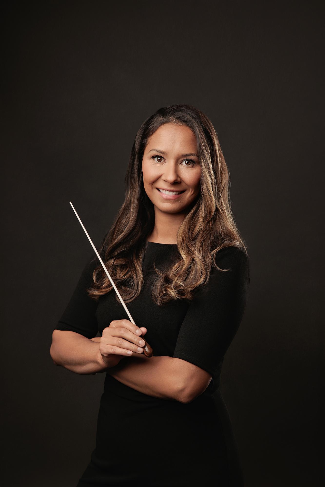 Unique personal branding photography Calgary: Female conductor in a black dress holding a baton at an angle with a confident smile against a dark backdrop