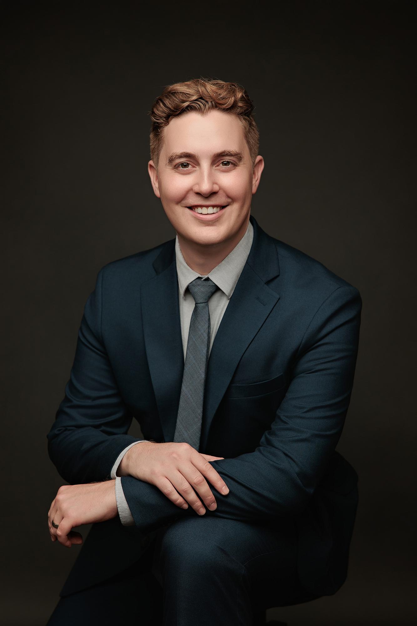 Executive headshot Calgary studio: Professional man in a navy suit and grey tie seated and leaning forward with hands clasped against a dark background
