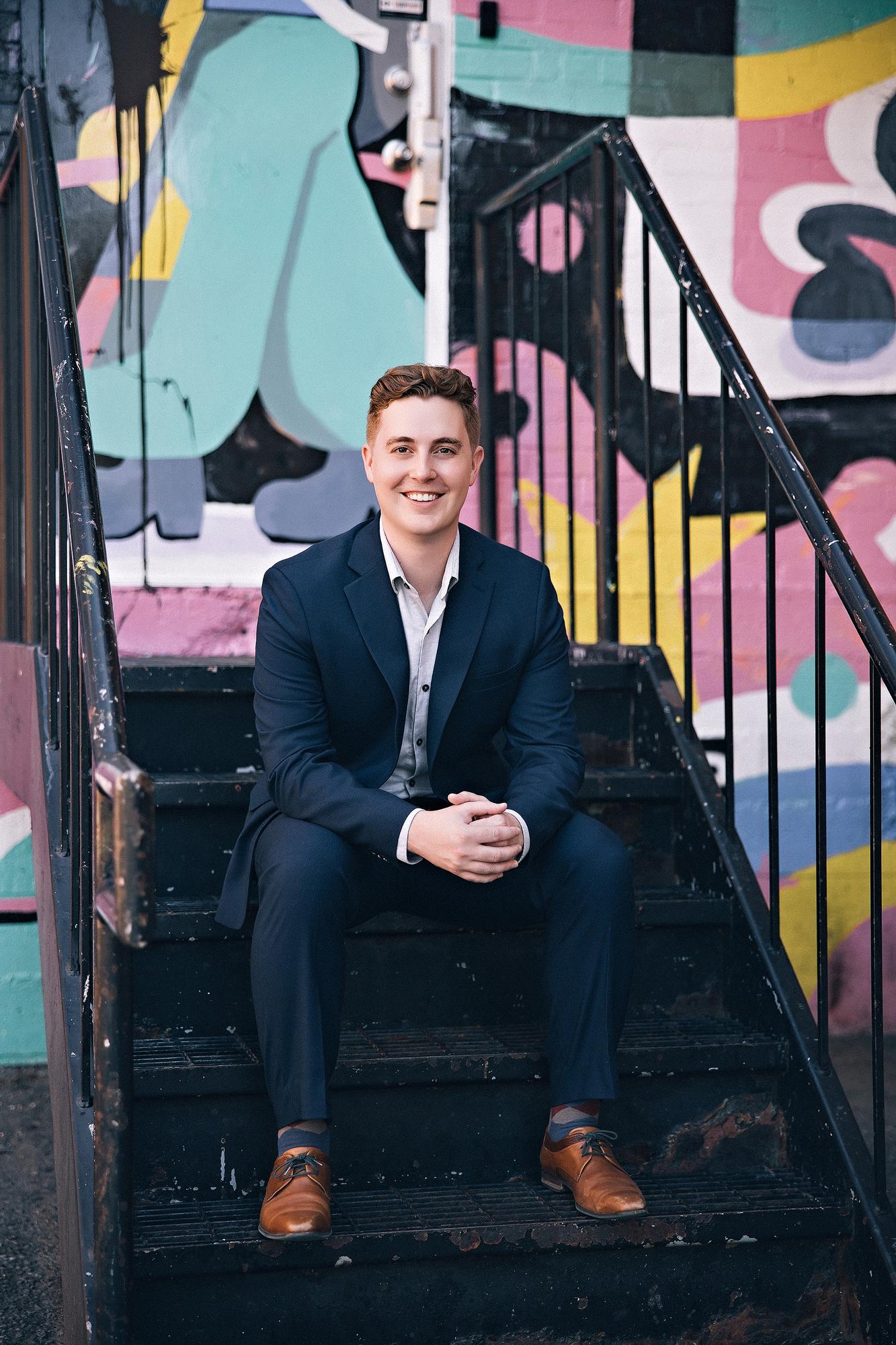 Outdoor professional headshot Calgary: Smiling man in a navy suit seated on stairs in front of a vibrant colourful street mural in Calgary