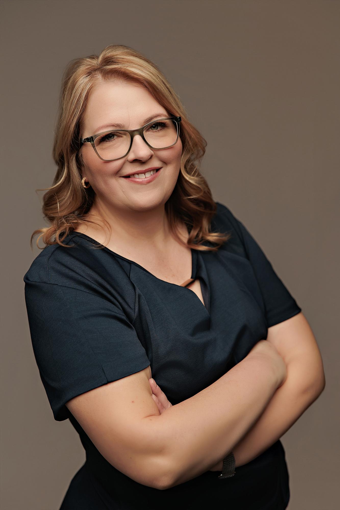 Corporate headshot photography Calgary: Confident woman with glasses in a dark teal short-sleeve top with arms crossed and a natural smile against a warm taupe background