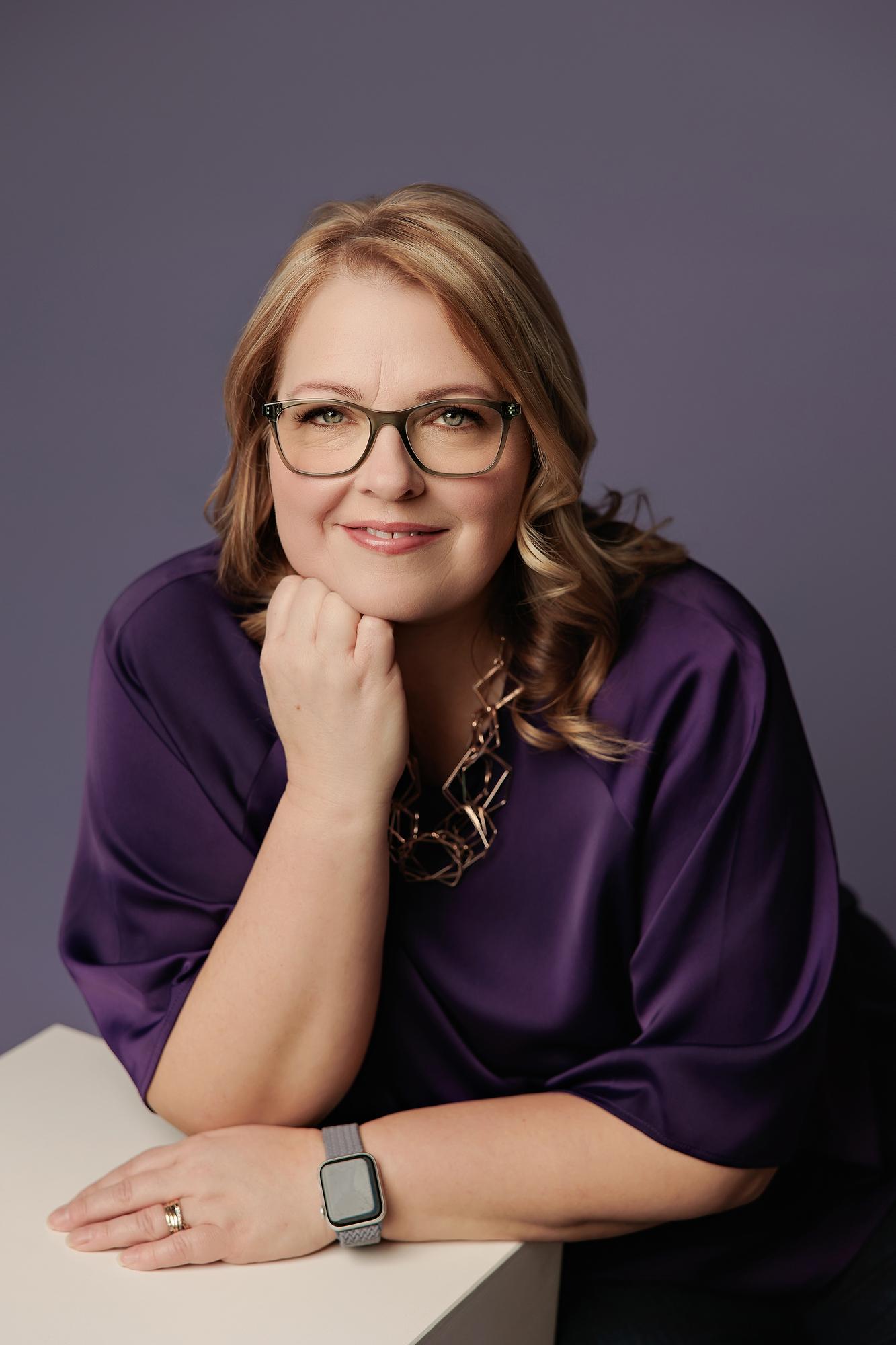 Professional headshot Calgary: Approachable woman with glasses in a purple silk top resting her chin on her hand against a matching purple studio background