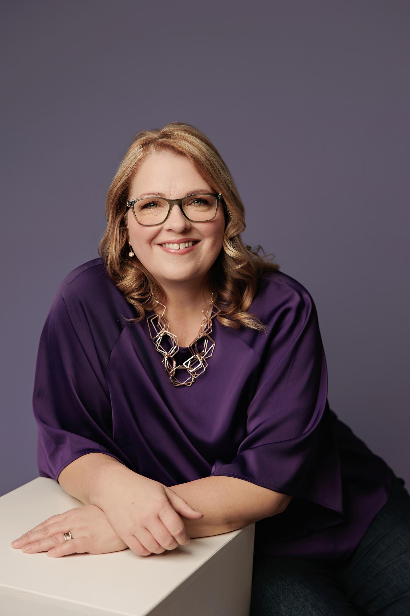 Corporate headshot Calgary studio: Smiling woman with glasses in a purple silk top leaning on a white cube with a gold statement necklace, purple background