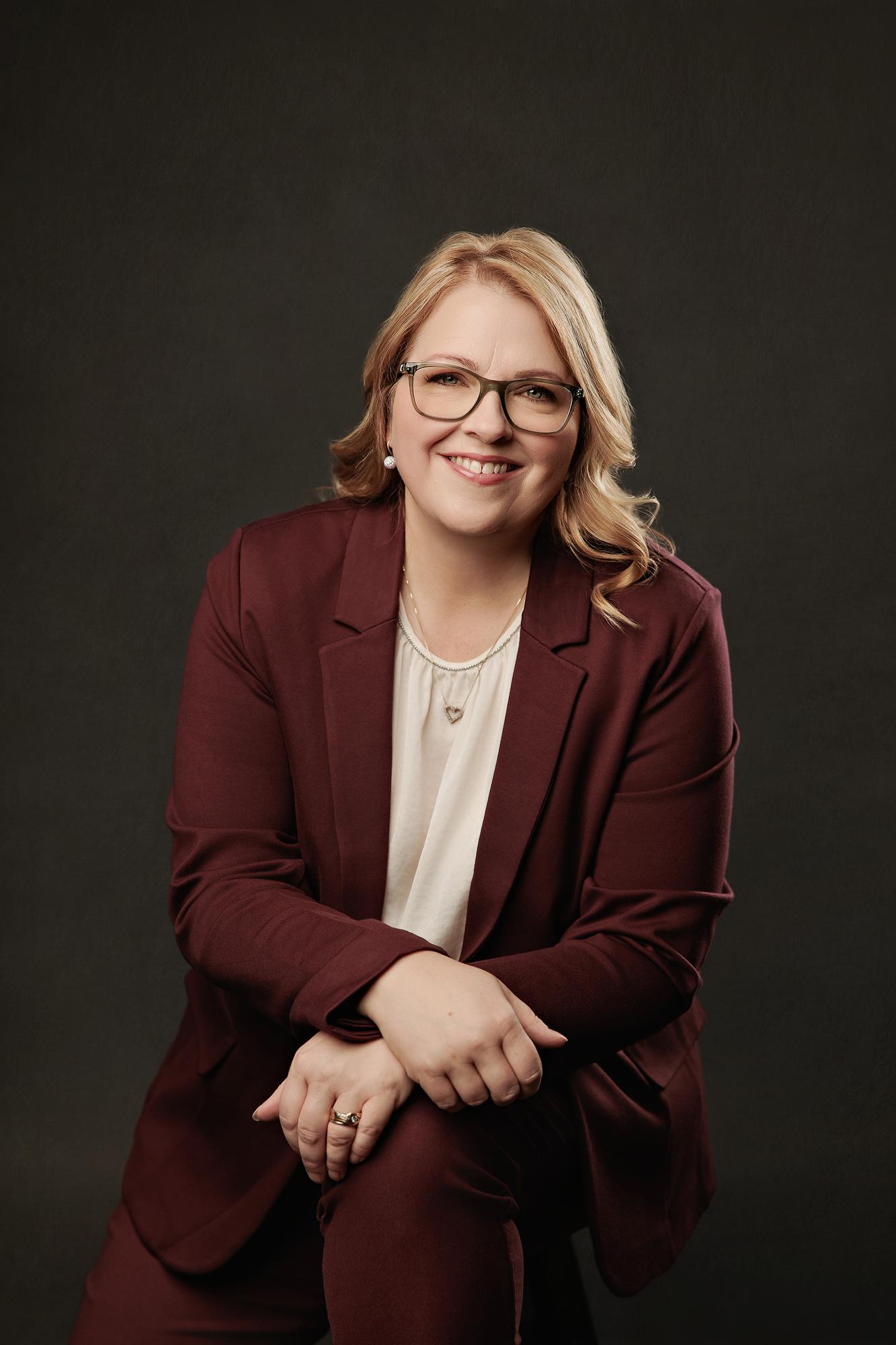 Executive headshot photography Calgary: Confident woman with glasses in a burgundy blazer seated with hands clasped and a warm smile against a dark background
