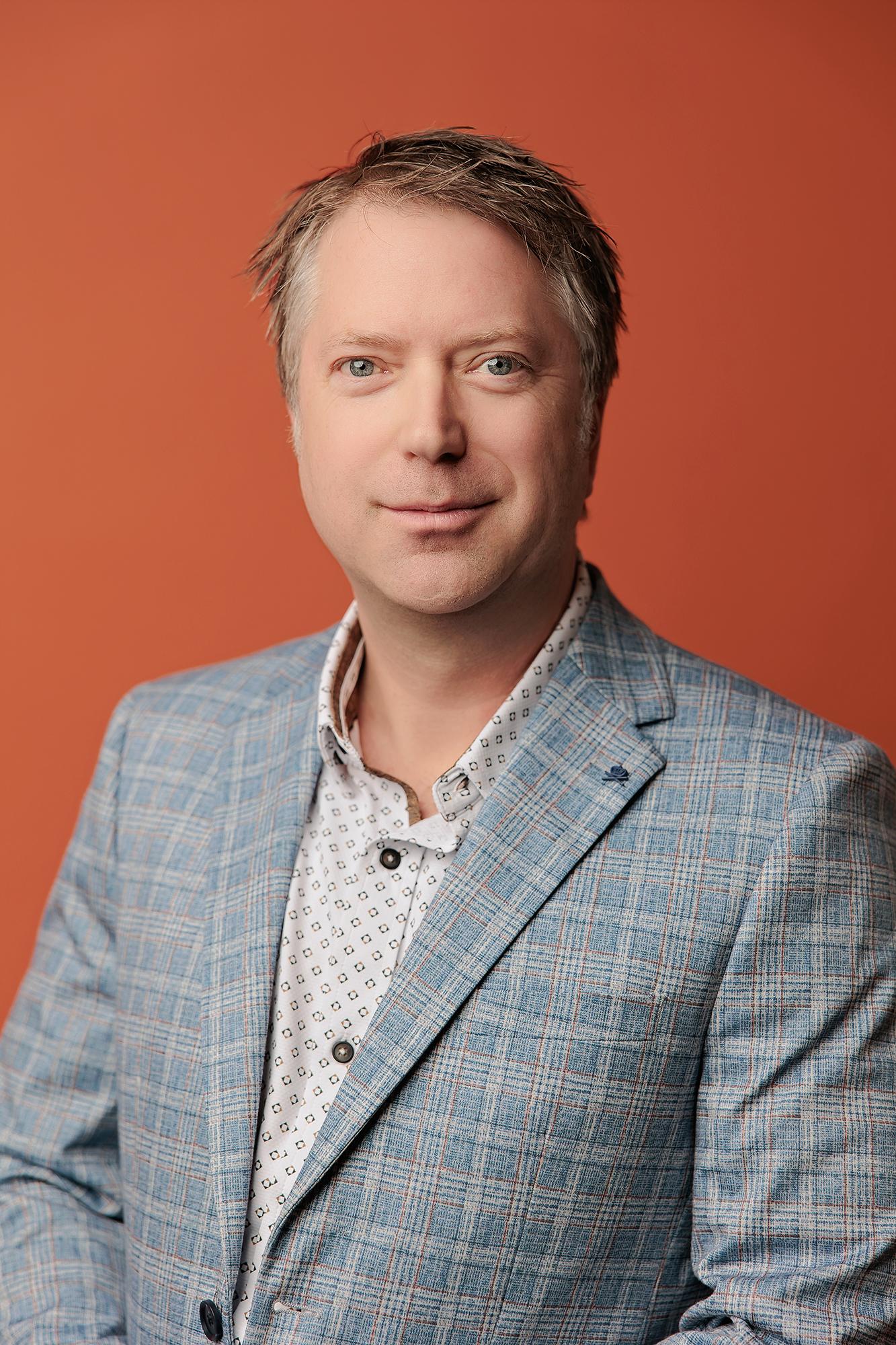 Professional headshot Calgary studio: Approachable male professional with blue eyes in a light blue plaid blazer and white geometric dot-print shirt with a relaxed expression against a bold orange background