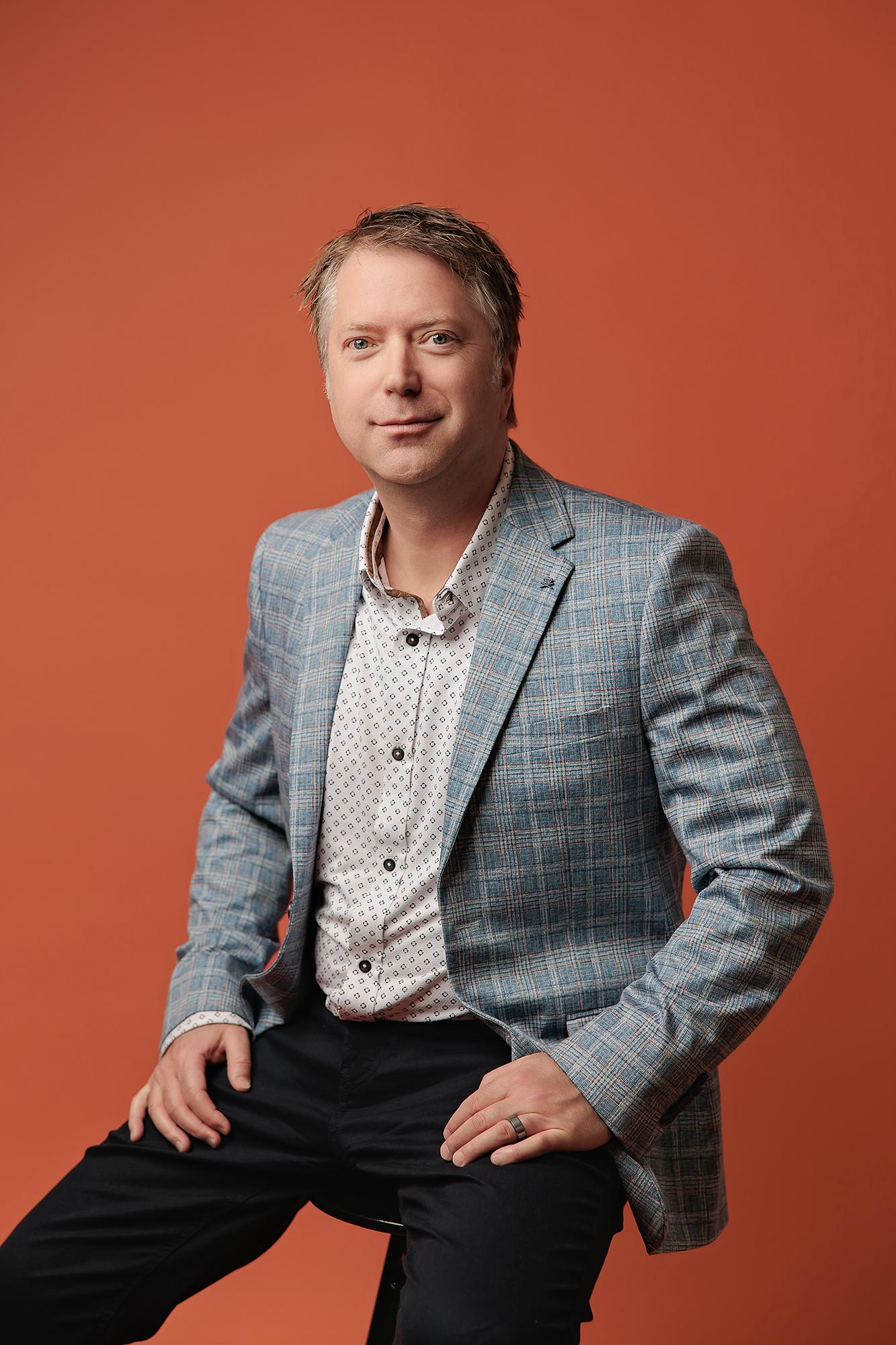 Professional headshot Calgary studio: Confident male professional in a light blue plaid blazer and white dot-print shirt seated on a stool with hands on knees and a direct gaze against a bold orange background