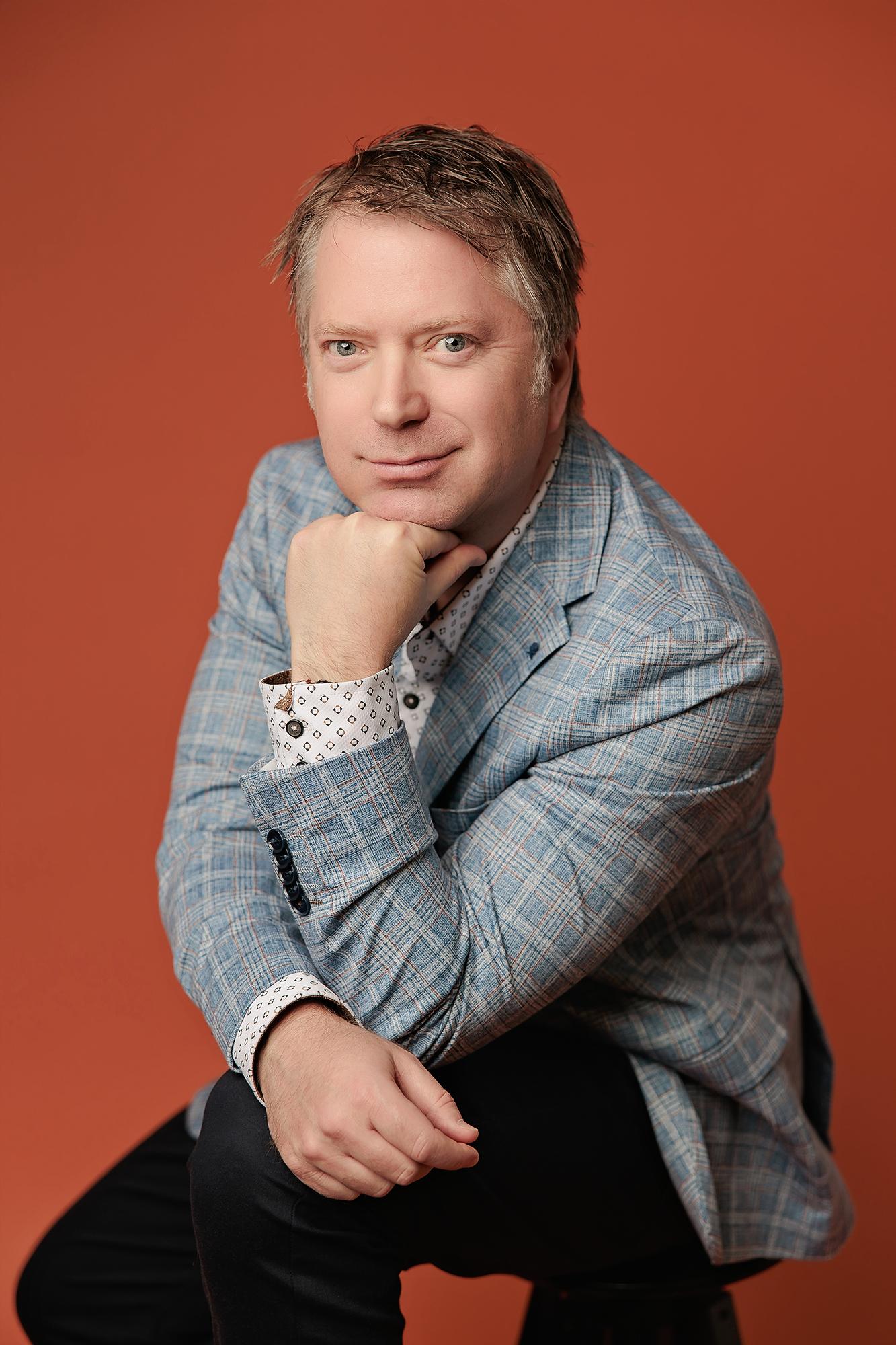 Professional headshot Calgary studio: Charismatic male professional in a light blue plaid blazer seated and leaning forward with chin resting on fist and a composed expression against a bold orange background