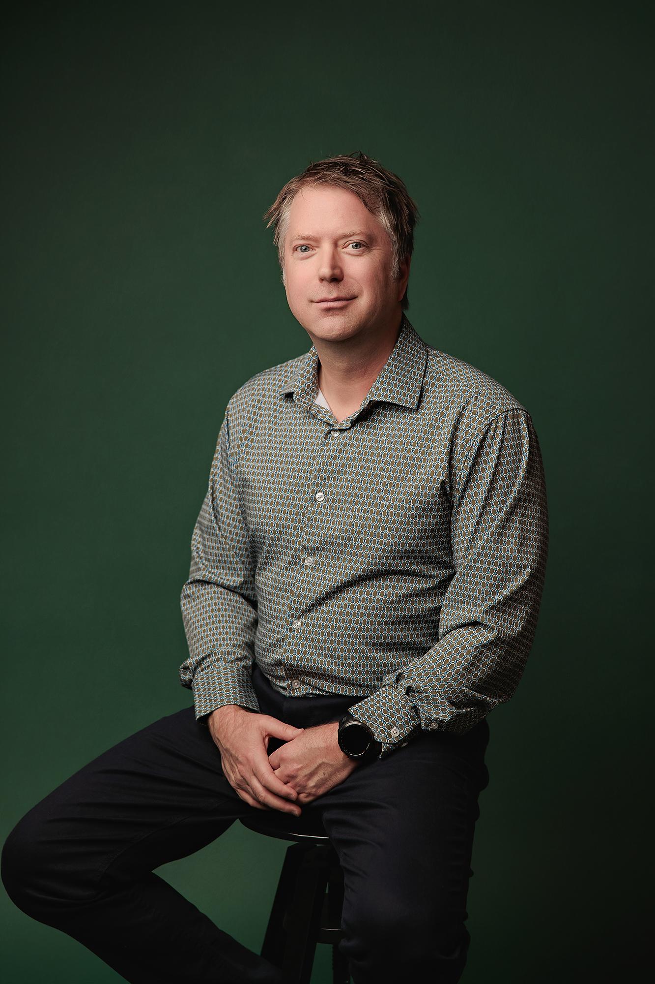 Professional headshot Calgary studio: Relaxed male professional in a teal-green geometric dot-print shirt seated on a stool with hands clasped and a subtle smile against a rich emerald green background