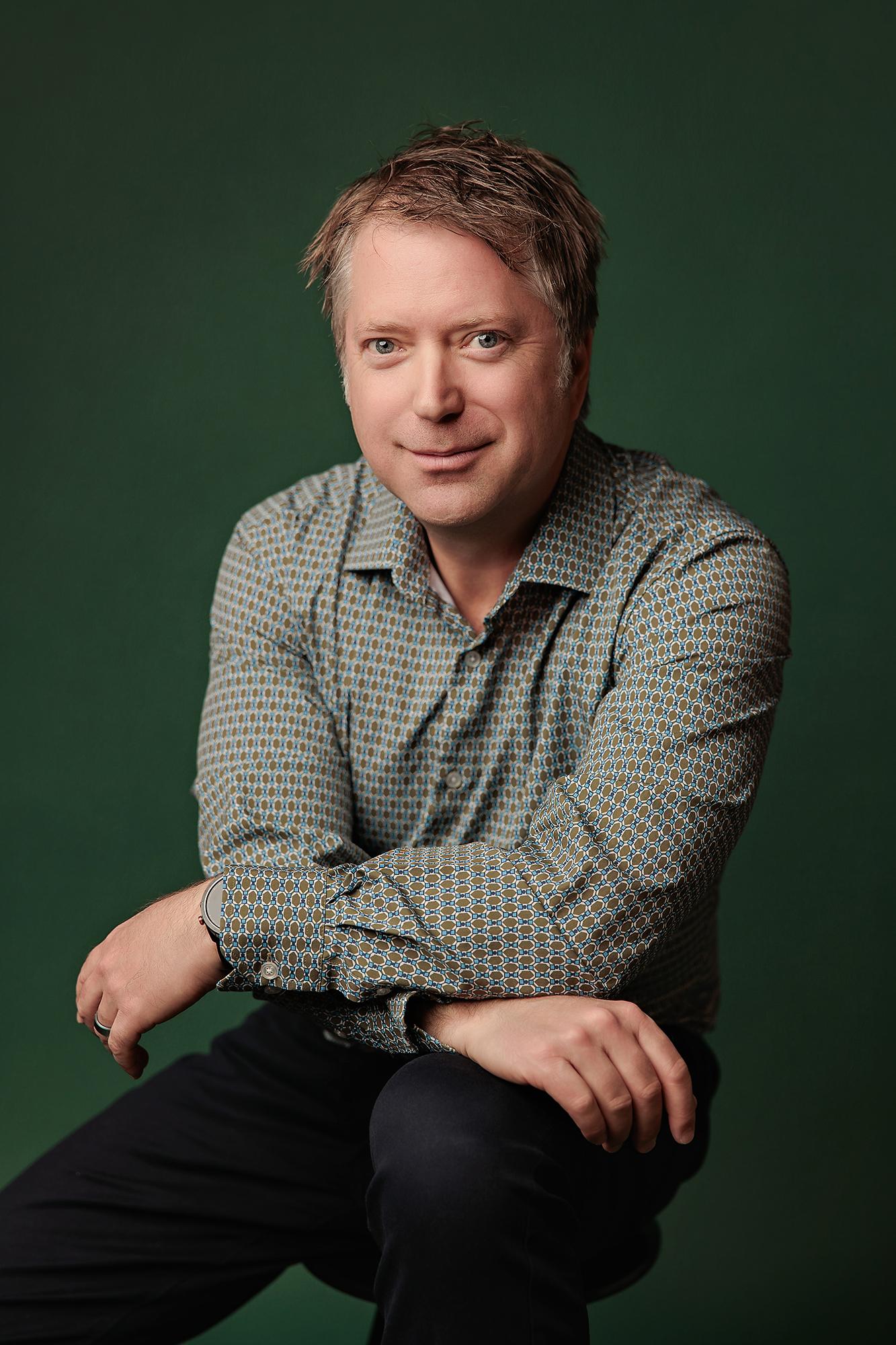 Professional headshot Calgary studio: Confident male professional in a teal-green geometric dot-print shirt seated and leaning forward with arms resting on knees and a direct gaze against a rich emerald green background