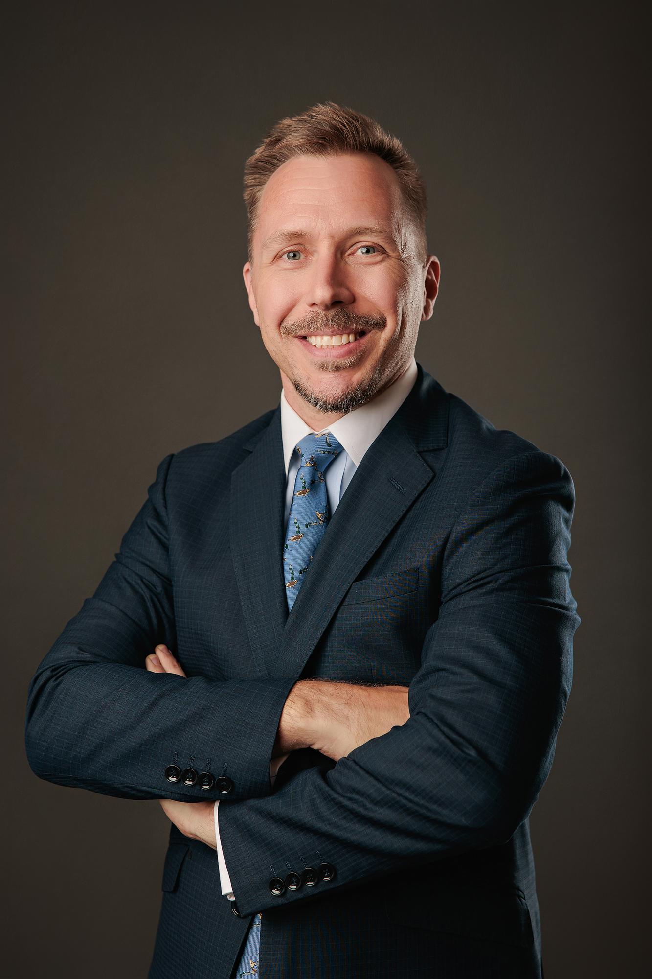 Professional headshot Calgary studio: Confident male executive with a goatee in a dark navy suit and blue novelty-print tie with arms crossed and a broad smile against a warm charcoal background