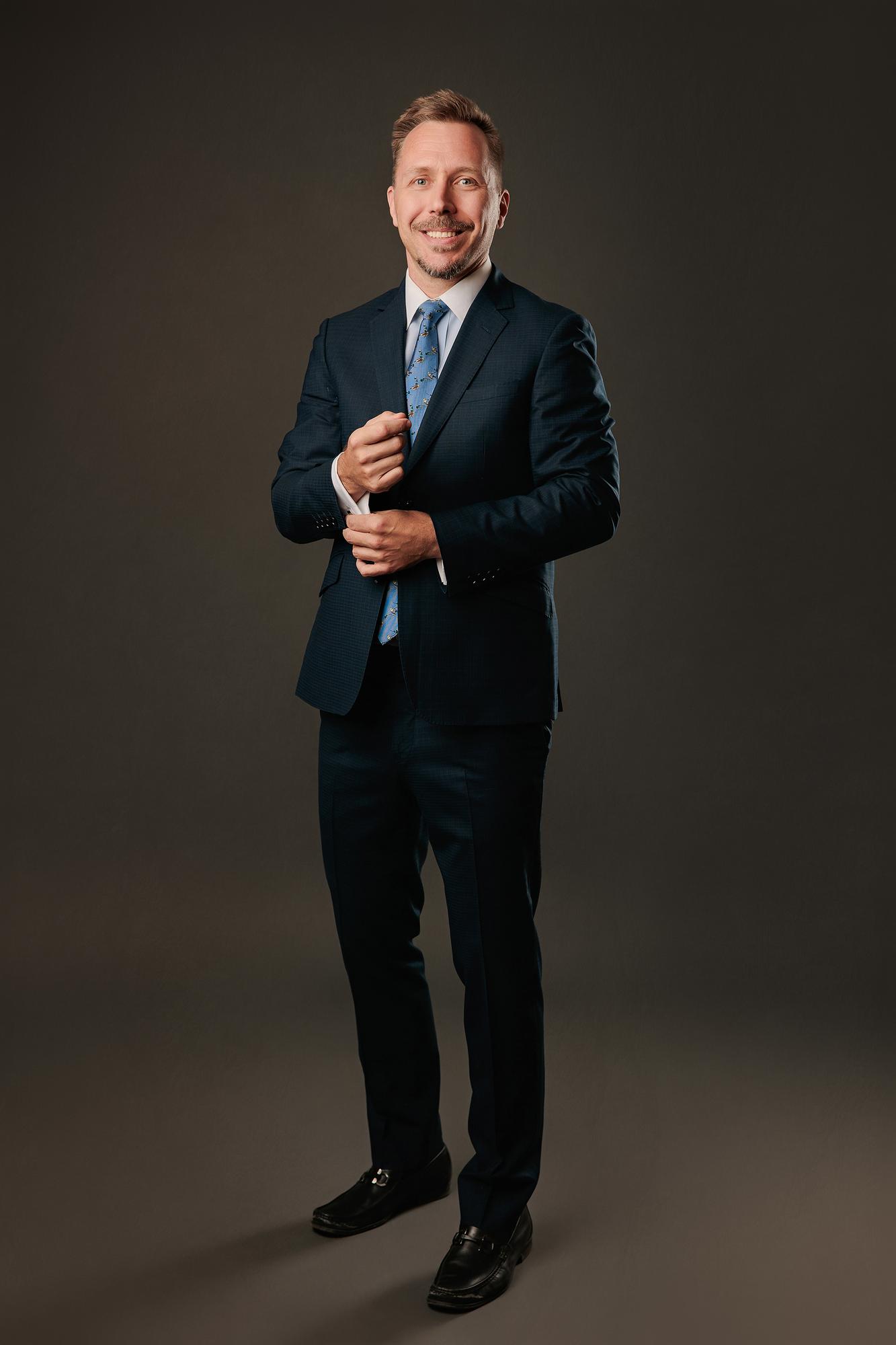 Full-length professional headshot Calgary studio: Polished male executive in a dark navy suit, white dress shirt, and blue novelty-print tie adjusting his cufflink with a warm smile and black leather loafers, charcoal background