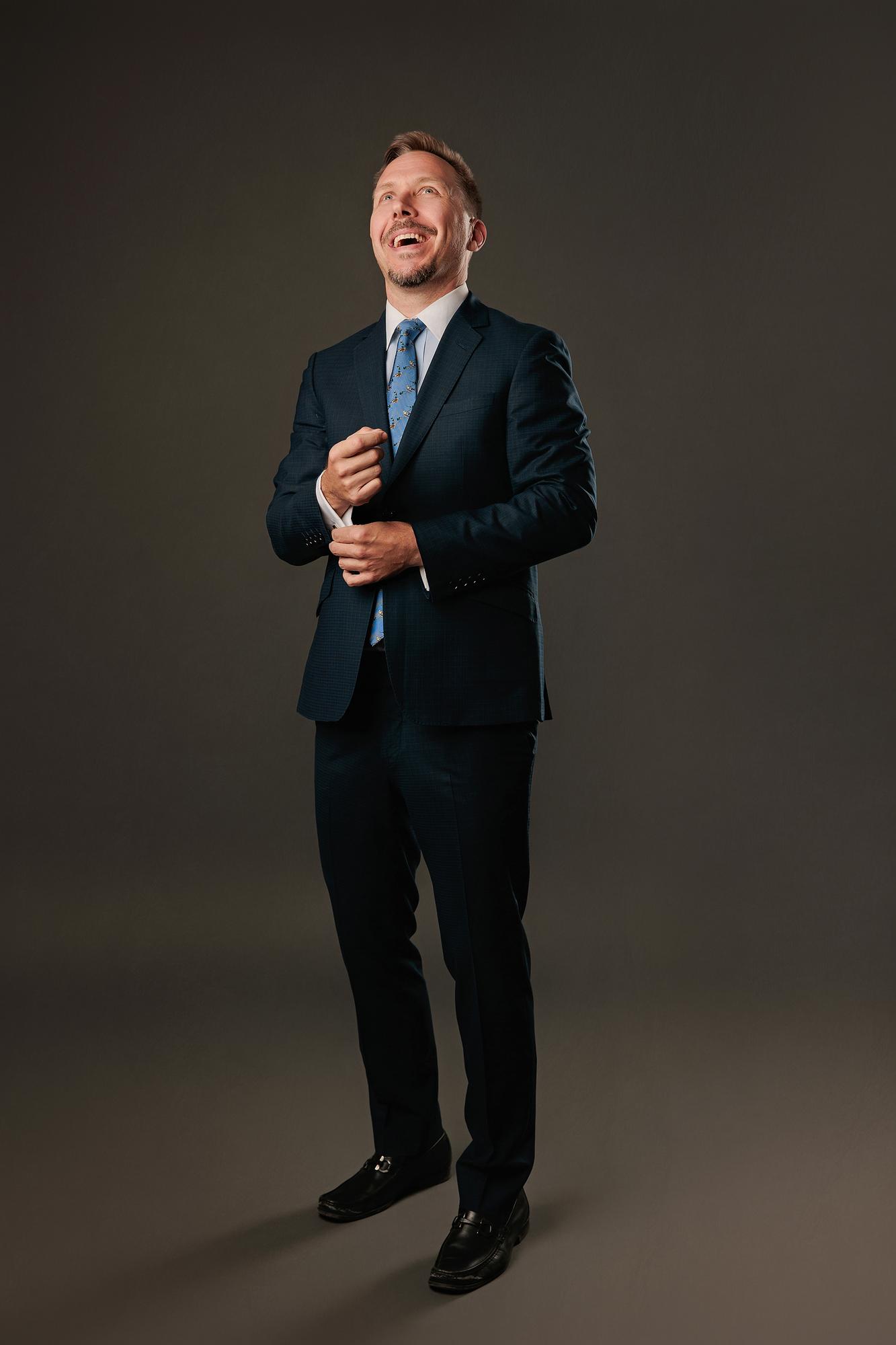 Full-length professional headshot Calgary studio: Joyful male executive in a dark navy suit and blue novelty-print tie laughing upward while adjusting his cufflink, black leather loafers, warm charcoal background