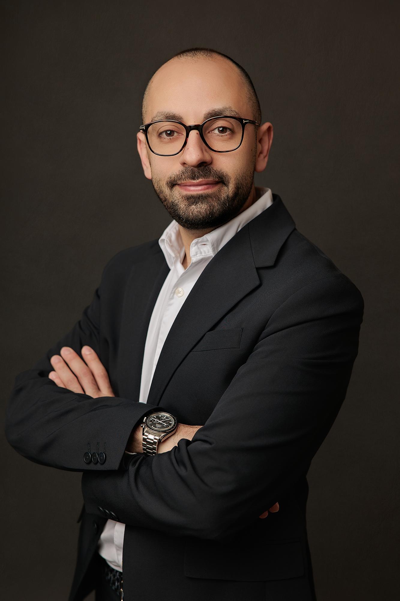 A confident man in a dark suit and glasses with crossed arms smiles subtly, a professional headshot captured by a Calgary headshot photographer.