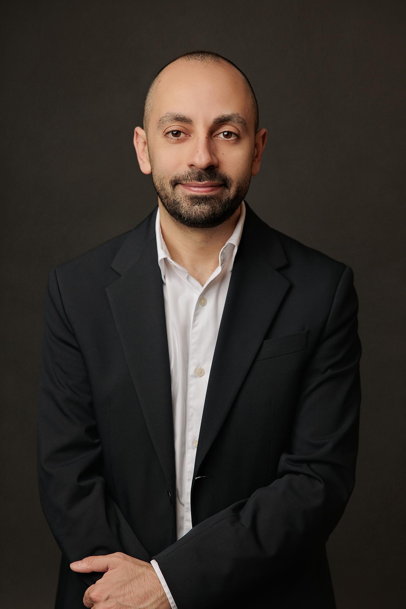 A professional Calgary headshot captures a confident man in a dark suit, hands clasped, smiling subtly at the camera against a dark studio background.