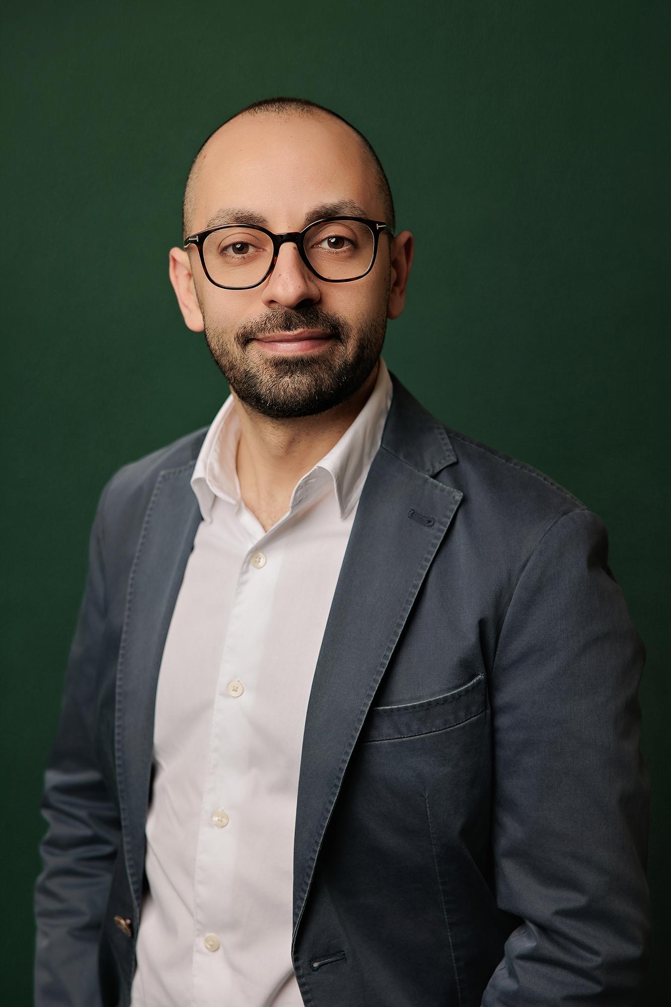 Calgary headshot photographer captures a smiling man with glasses and beard in a blue blazer against a dark green studio background.